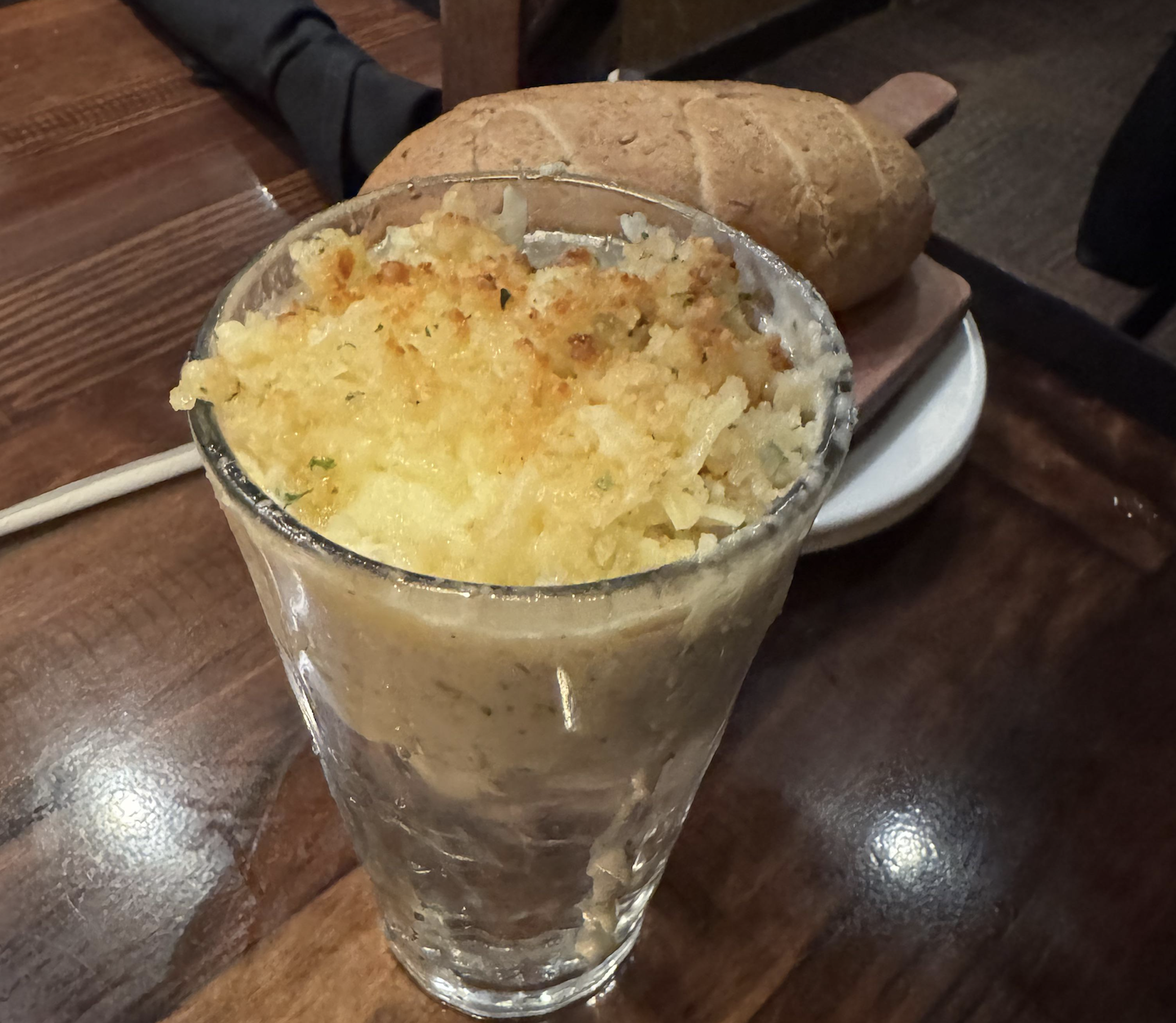 A glass filled with a baked dish topped with crispy shredded potatoes, placed on a wooden table next to a loaf of bread on a small cutting board.
