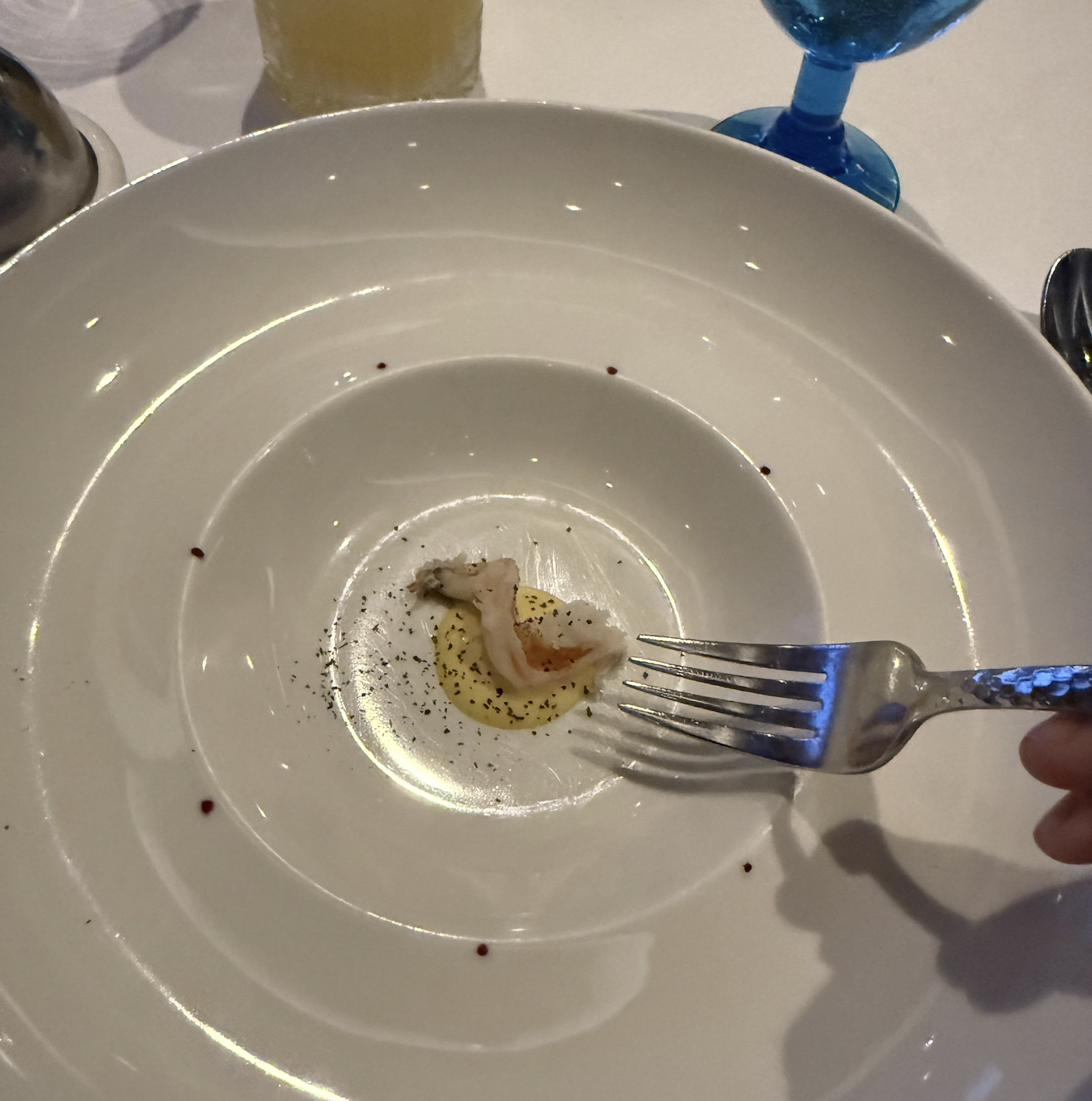 A mostly empty white plate with a small piece of food in the center, some sauce, and garnish. A hand holds a fork, about to pick up the food. Blue glass and drink are in the background.