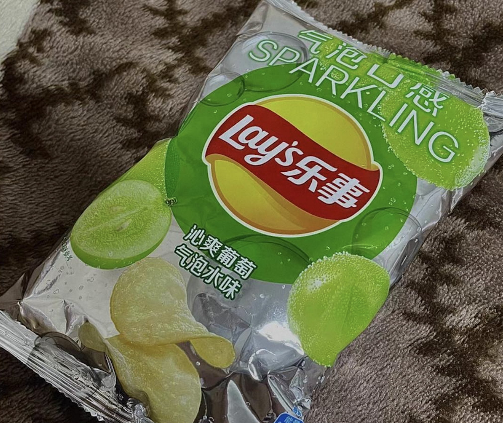A bag of Lay’s sparkling grape-flavored potato chips with green grape graphics lies on a brown patterned blanket.