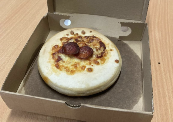 A small, plain pizza with two pepperoni slices sits in an open cardboard box on a wooden table.