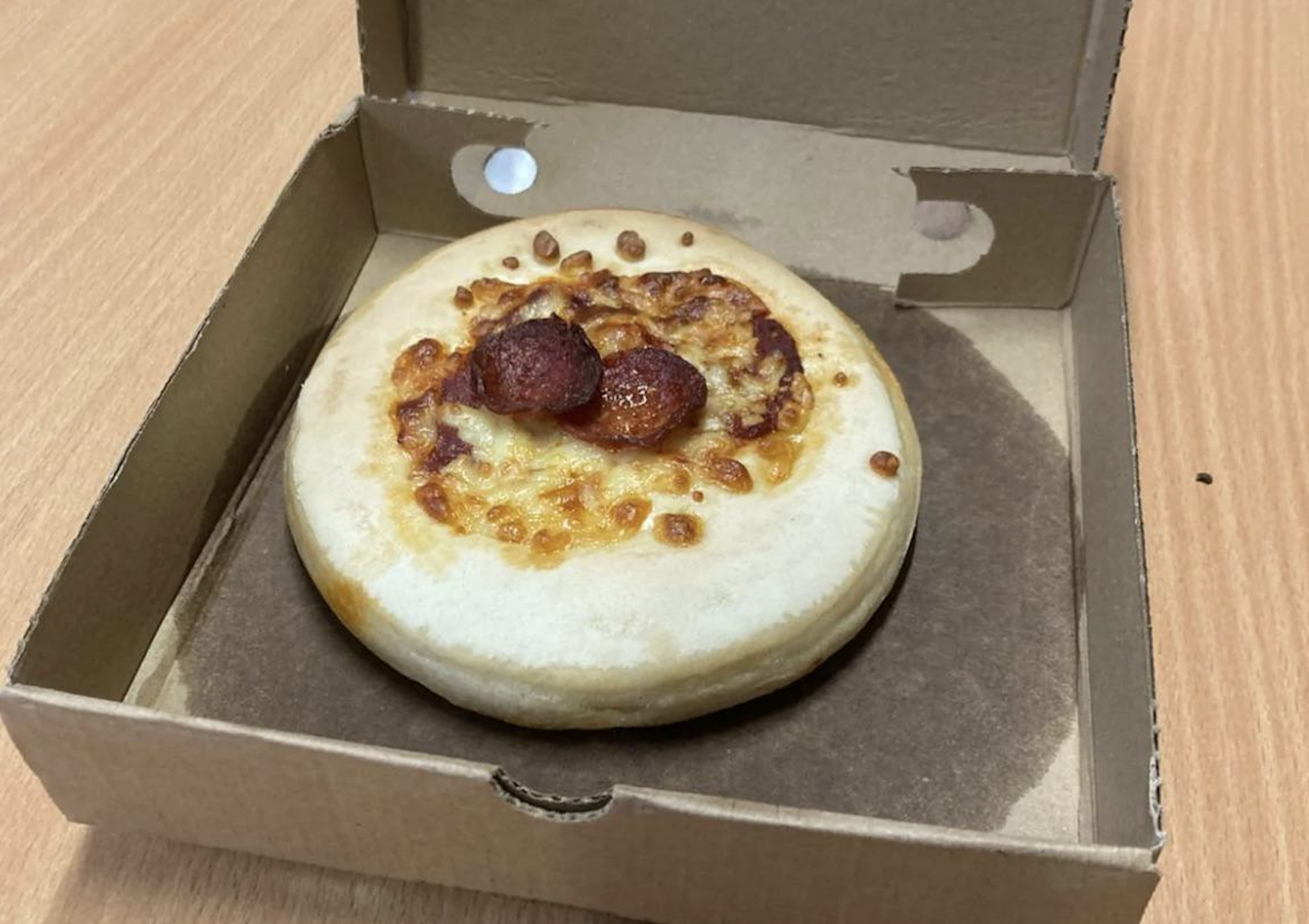 A small, plain pizza with two pepperoni slices sits in an open cardboard box on a wooden table.
