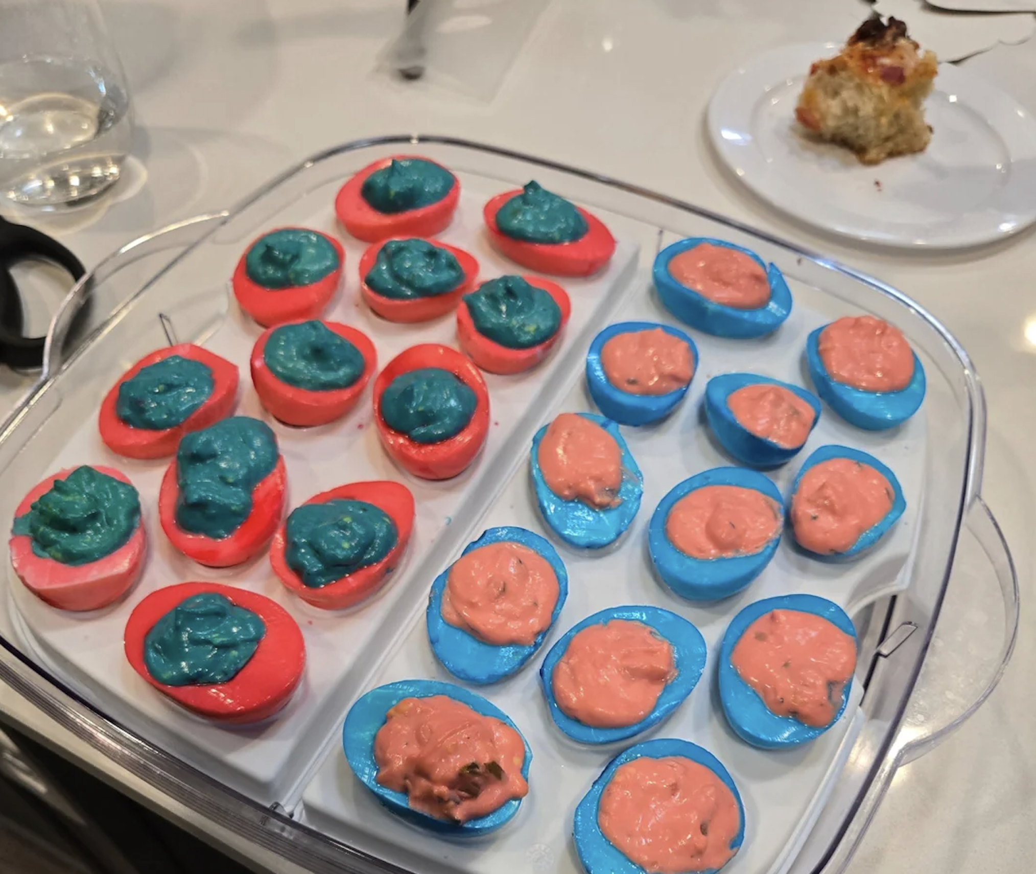 A tray of brightly colored deviled eggs, with red eggs filled with blue-green filling and blue eggs filled with pink filling. A plate with food and a glass of water are in the background.