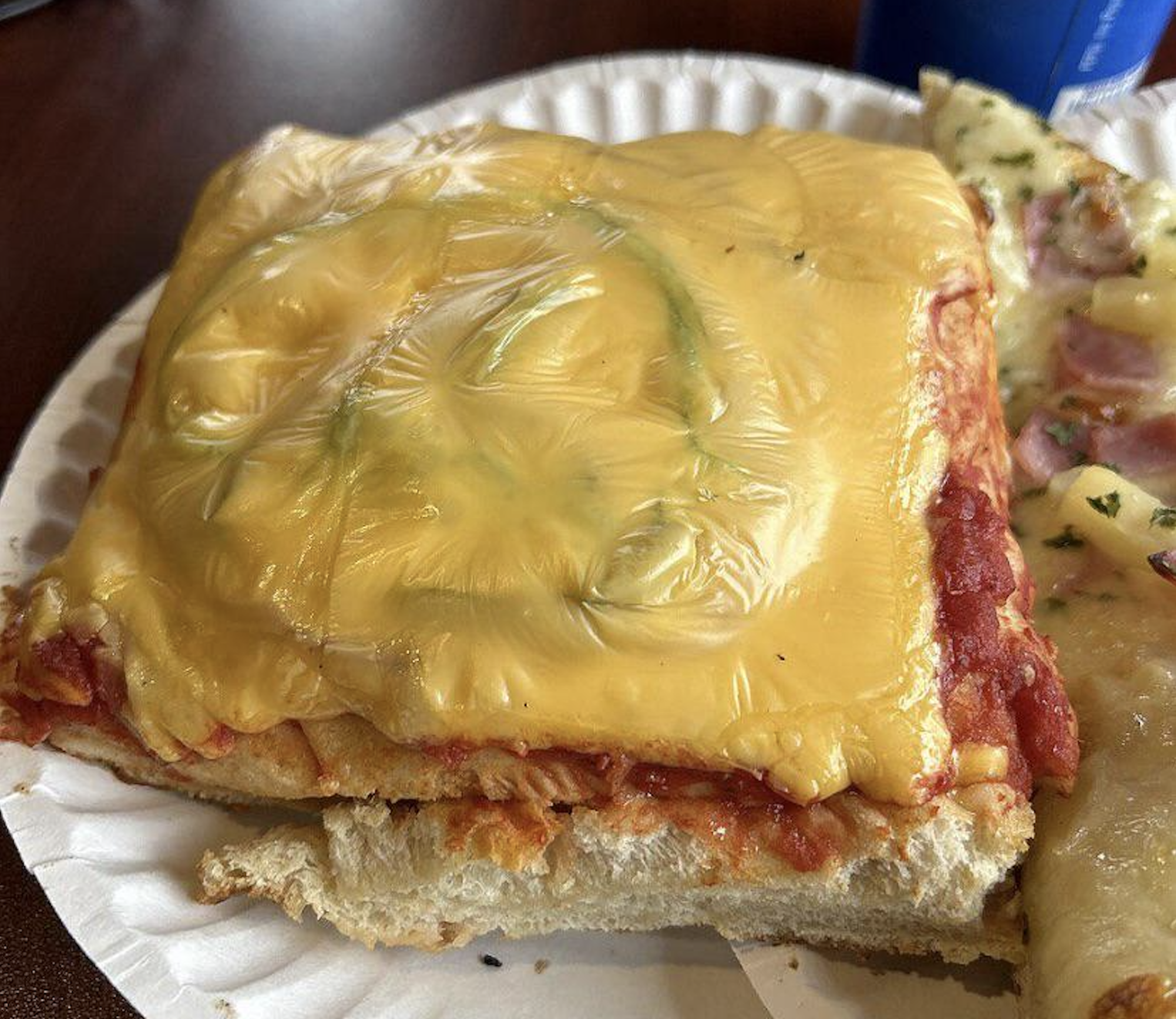 A slice of thick, rectangular pizza topped with tomato sauce and a melted slice of processed yellow cheese, served on a paper plate.