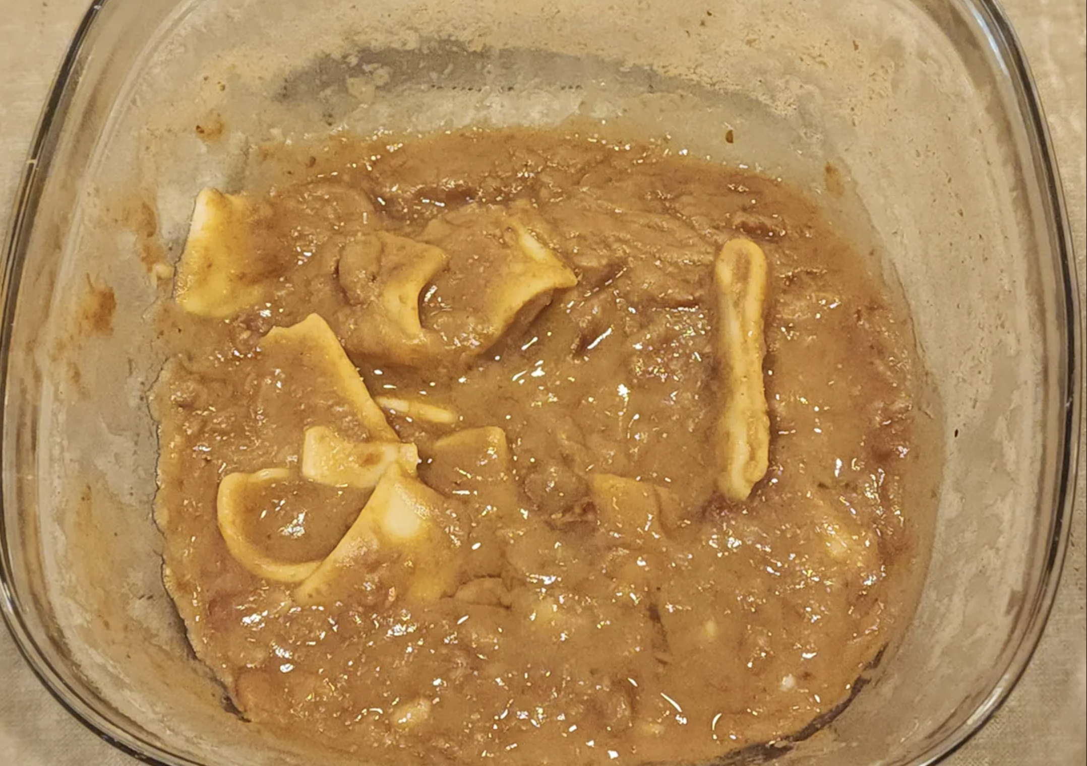 A glass dish contains a thick, brown stew with a few wide, flat pasta pieces partially submerged in the mixture. The surface is lumpy and glossy.