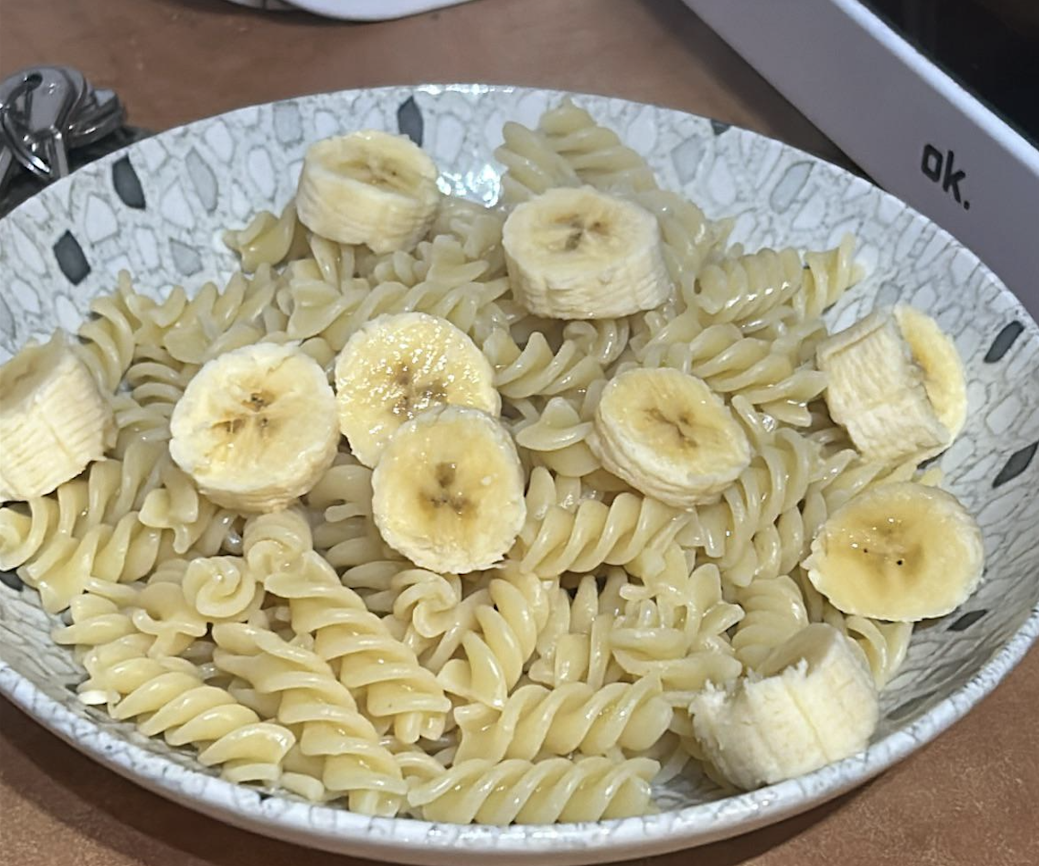A bowl of cooked rotini pasta topped with sliced bananas, placed on a patterned plate on a countertop.