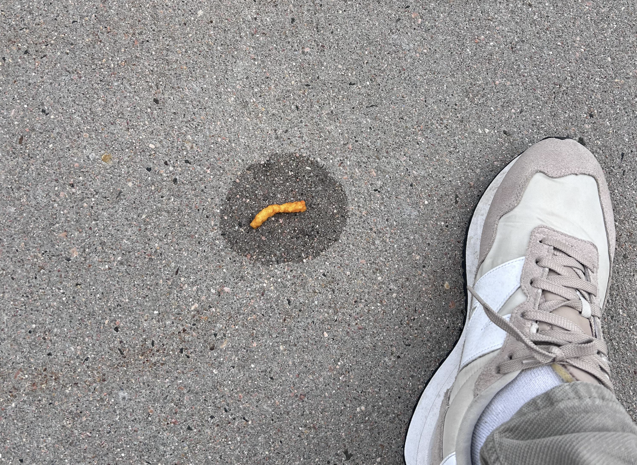 A yellowish worm-like object lies on gray pavement with a wet circle around it, next to a person’s beige and white sneaker.