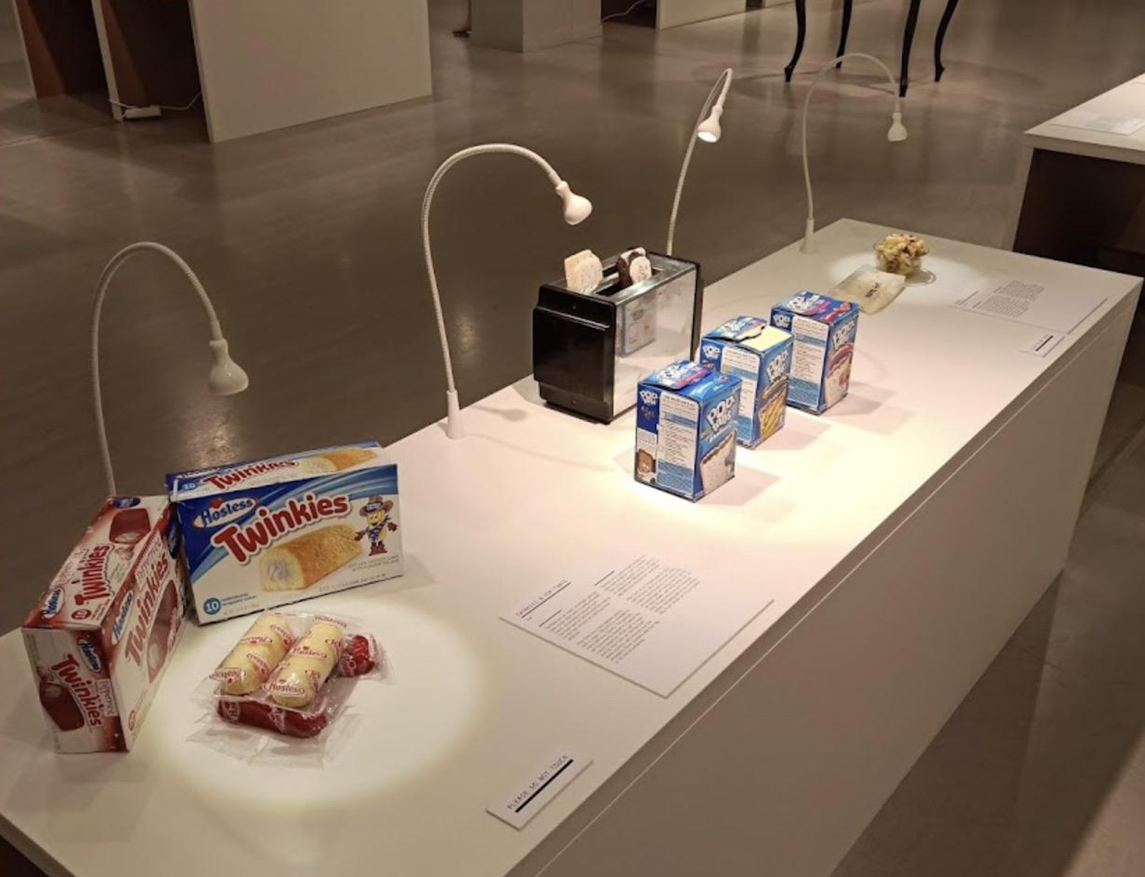 A museum display features Twinkies, cartons of milk, and other foods under small lights on a white platform, with information cards placed in front of each item.