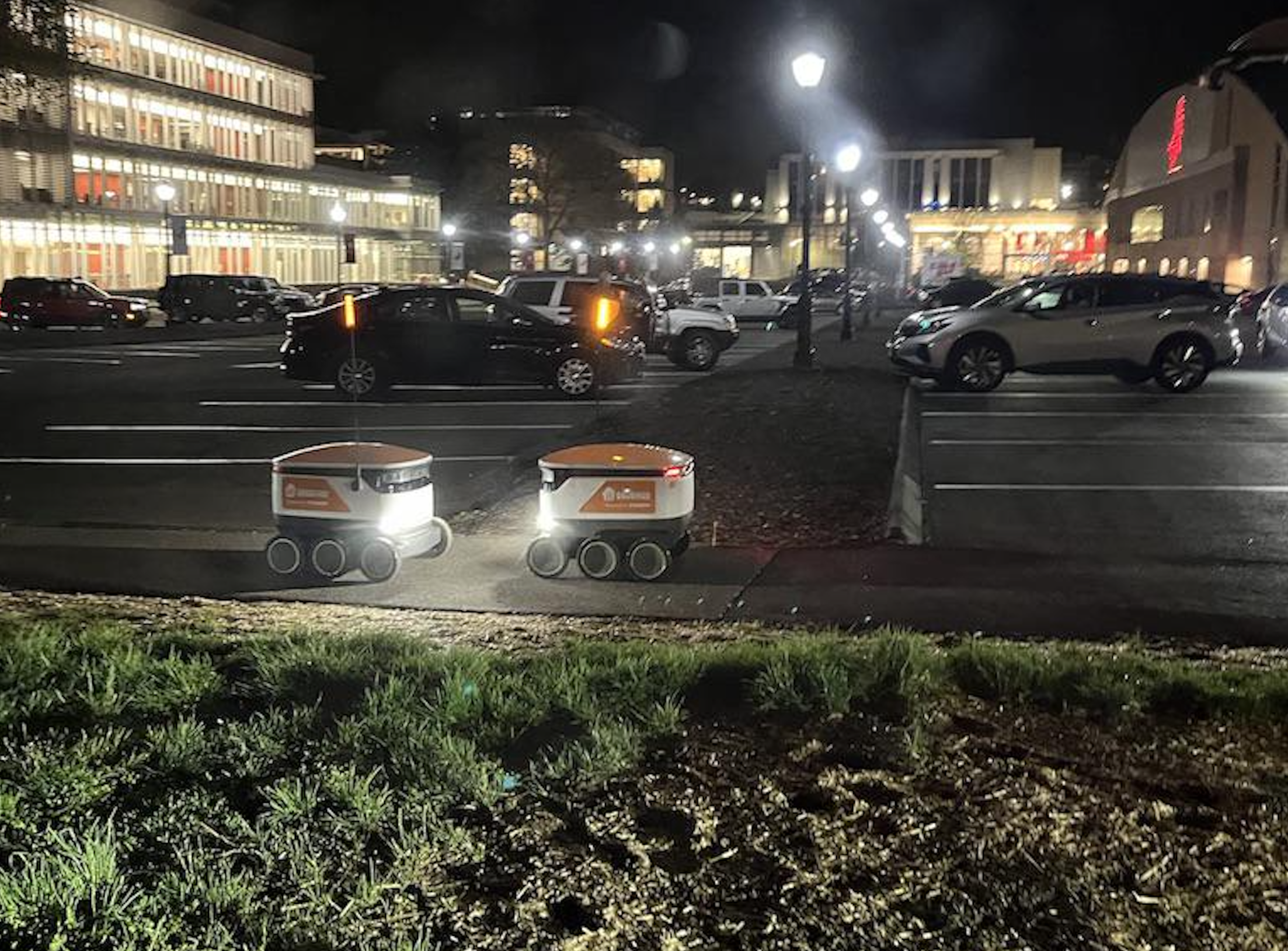 Two small delivery robots with headlights drive on a sidewalk at night beside a parking lot with parked cars and illuminated buildings in the background.