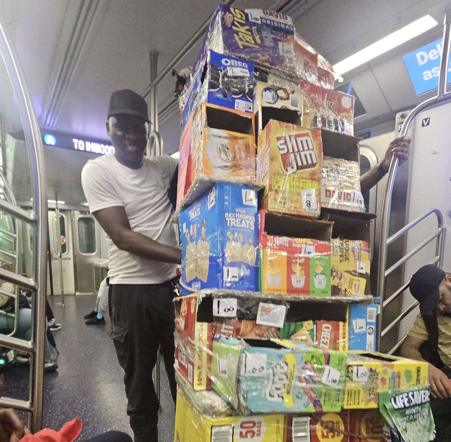 A person stands in a subway car, balancing a tall, stacked tower of colorful snack boxes, including Oreos, Slim Jims, and Lifesavers, with passengers seated around them.