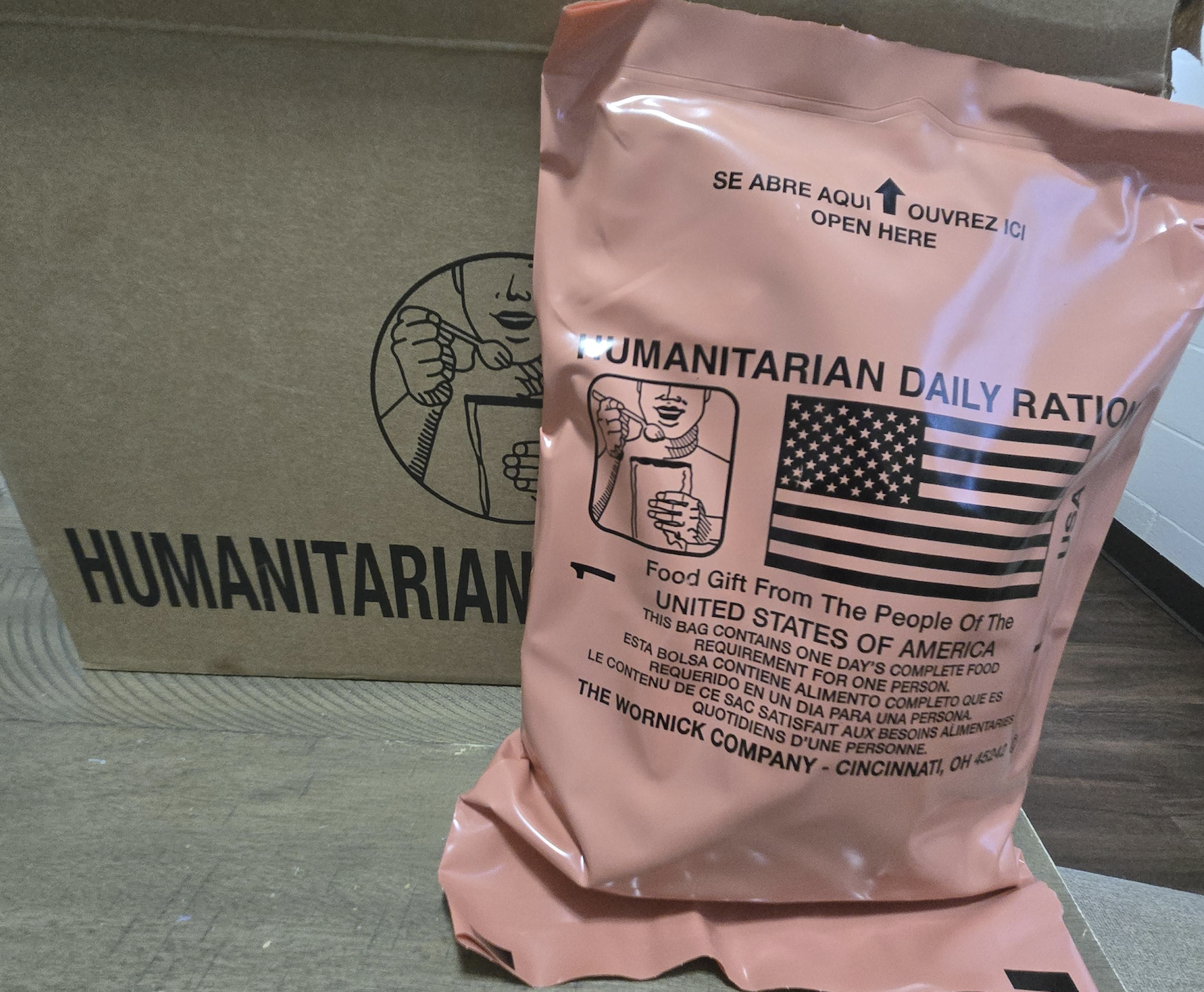 A brown box labeled “HUMANITARIAN” and a pink Humanitarian Daily Ration food pouch with American flag, instructions, and text indicating it is a food gift from the United States.