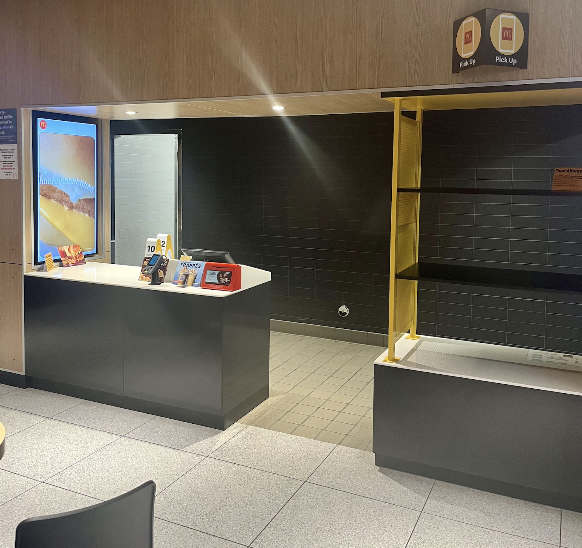 A modern fast-food restaurant counter with a digital menu, order pickup shelf, and minimal decor. The area is clean, with gray tiles and black walls.