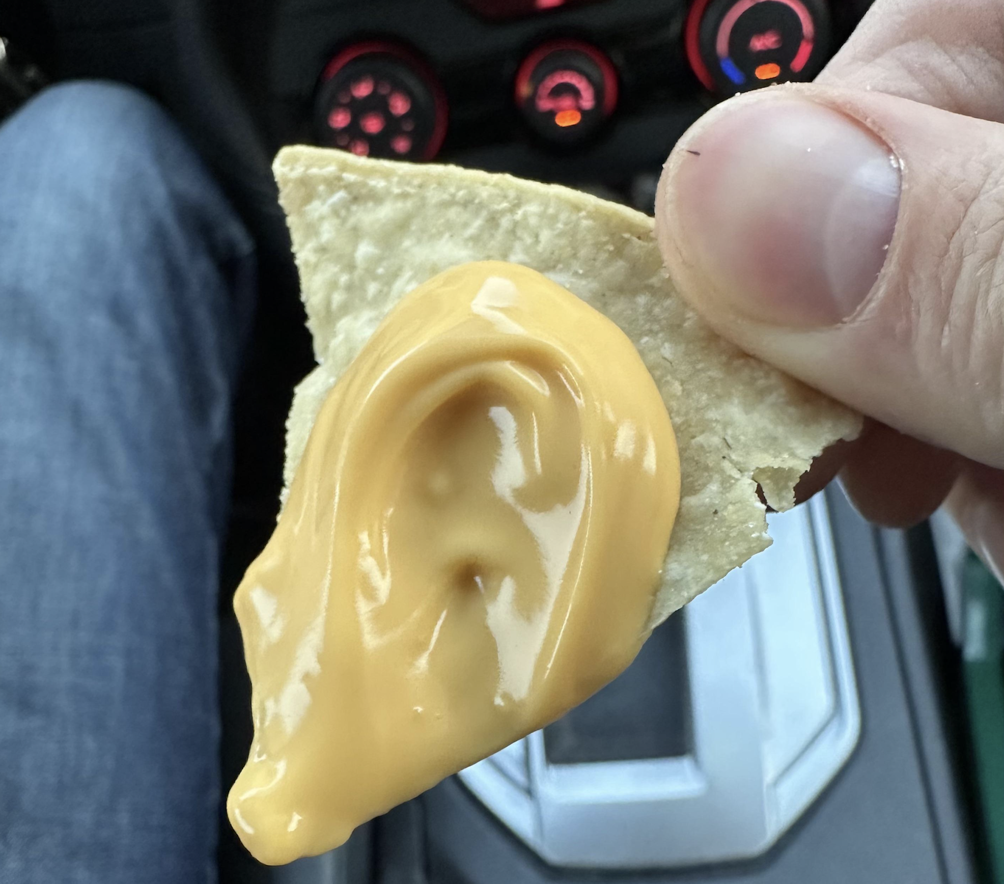 A hand holds a tortilla chip topped with nacho cheese, shaped like a human ear, inside a car with dashboard controls visible in the background.