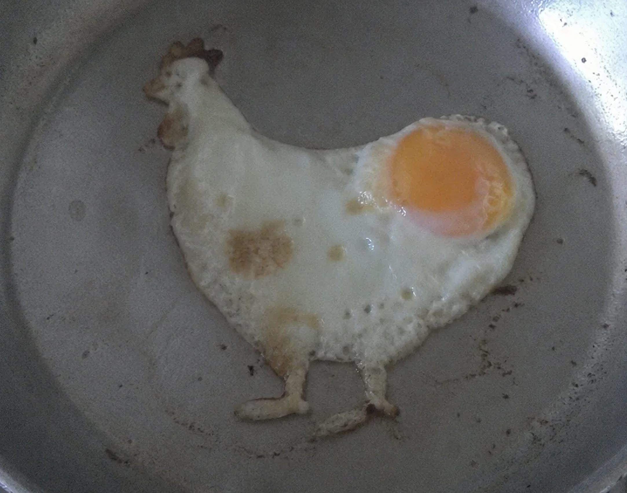 A fried egg in a pan is shaped like a chicken, with the egg yolk forming the body and white forming the head, beak, and legs.