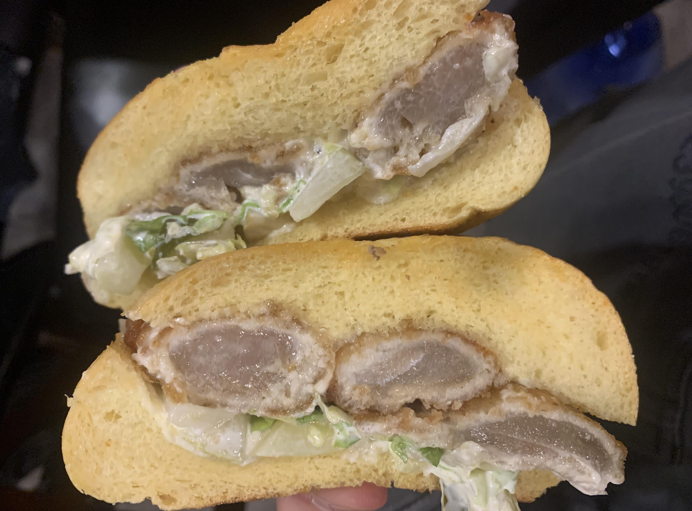 A sandwich cut in half, filled with breaded fried meat, lettuce, and creamy sauce, on a soft yellowish bun.