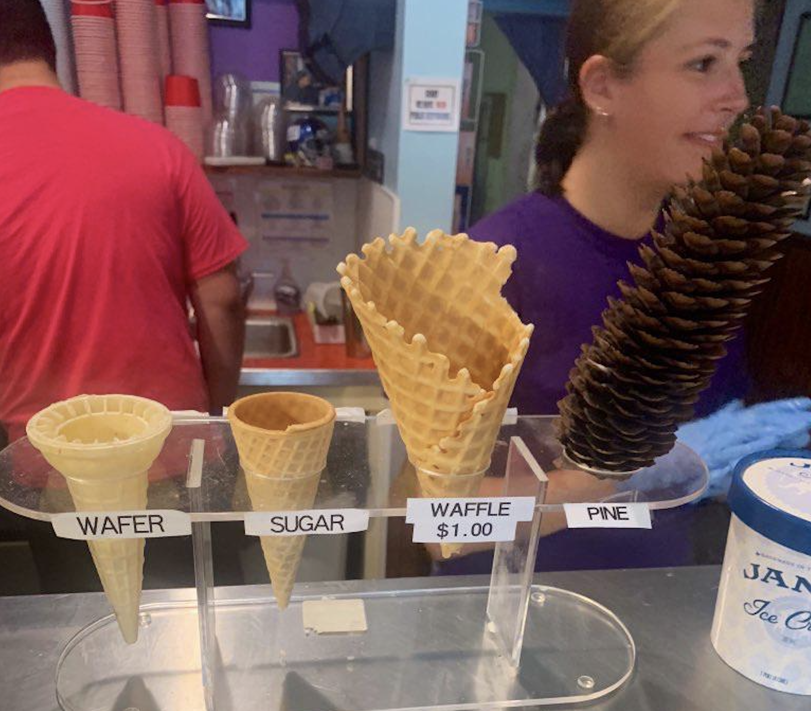Four items on display: wafer cone, sugar cone, waffle cone for $1, and a pine cone. Two people are in the background at an ice cream counter.