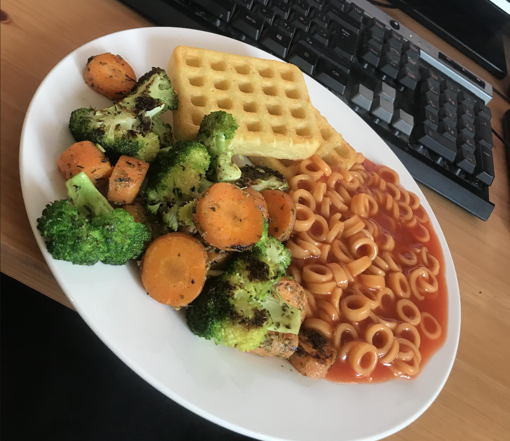 A plate with two potato waffles, baked beans with spaghetti hoops, and roasted broccoli and carrots, placed on a desk near a computer keyboard.