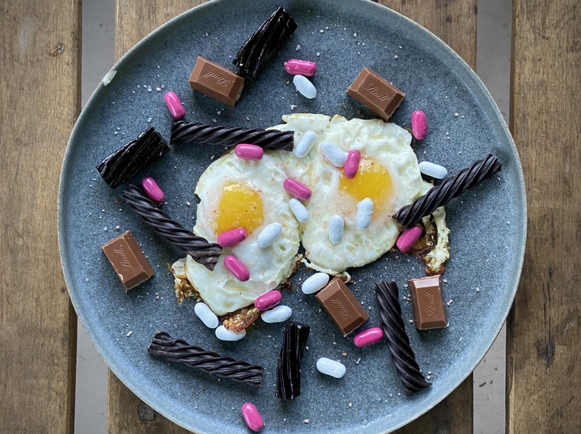 A blue plate with two fried eggs, chocolate pieces, black licorice sticks, and pink and blue capsules, set on a wooden surface.