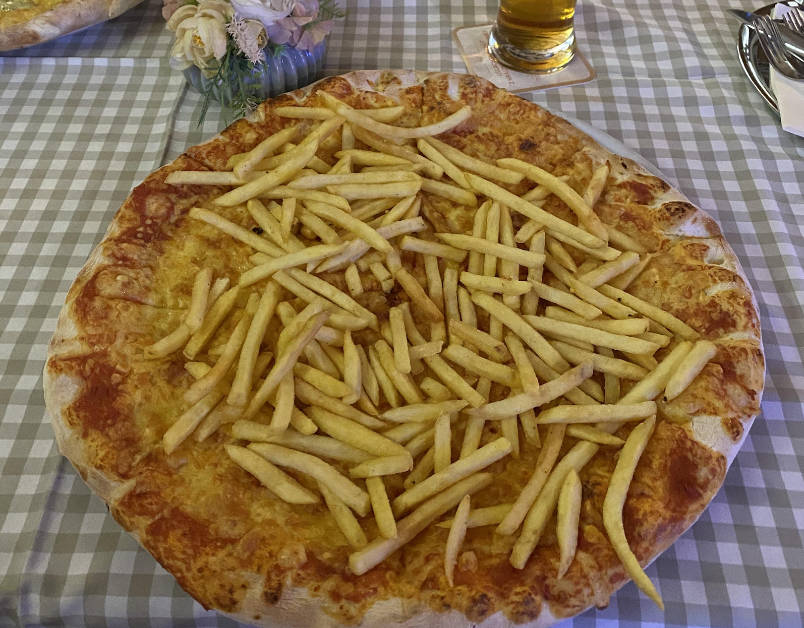 A large pizza topped with French fries sits on a checkered tablecloth, with a drink and a flower arrangement in the background.
