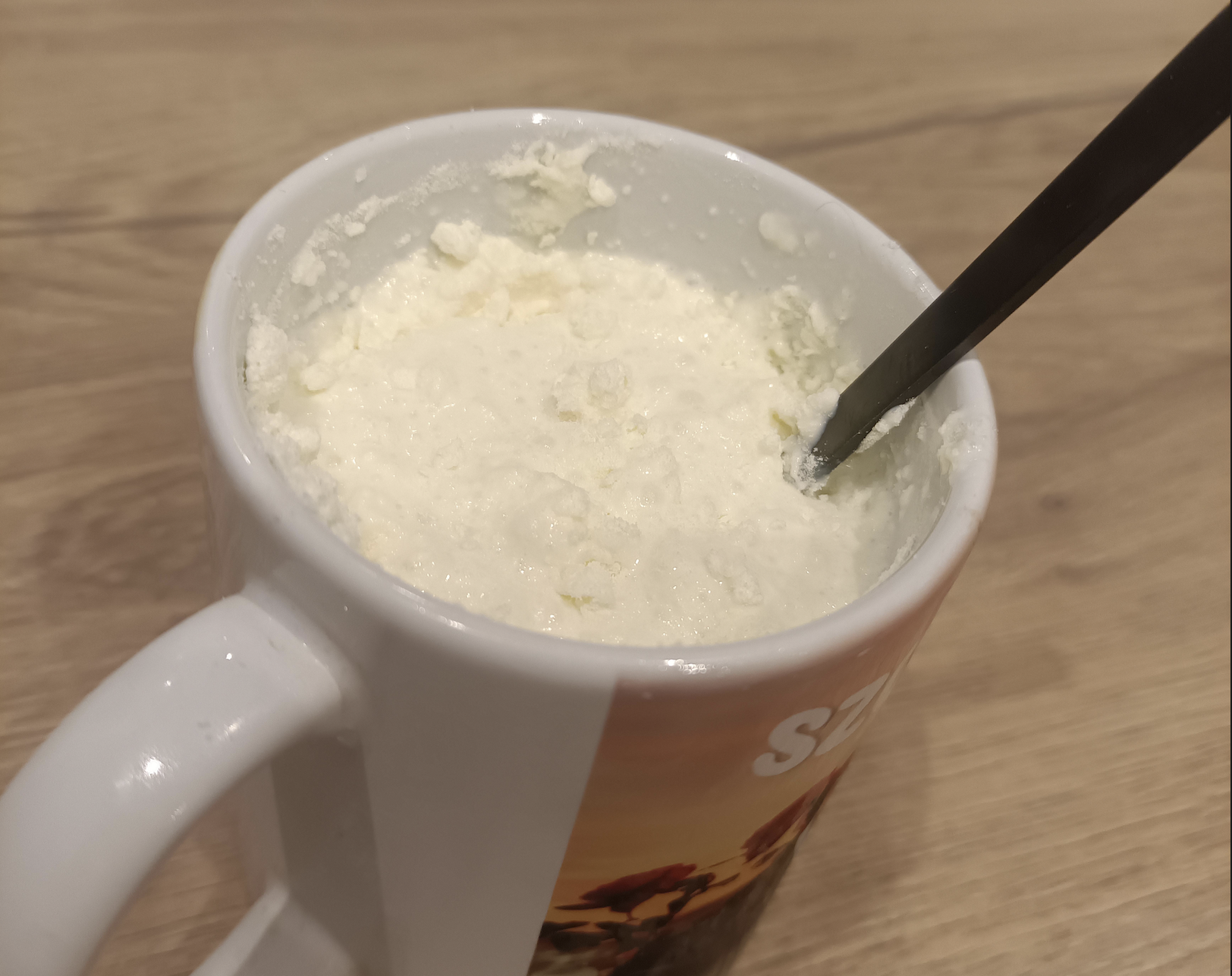 A white mug filled with a lumpy, creamy substance, possibly homemade cottage cheese or curd, sits on a wooden surface. A black spoon is inserted into the mixture.