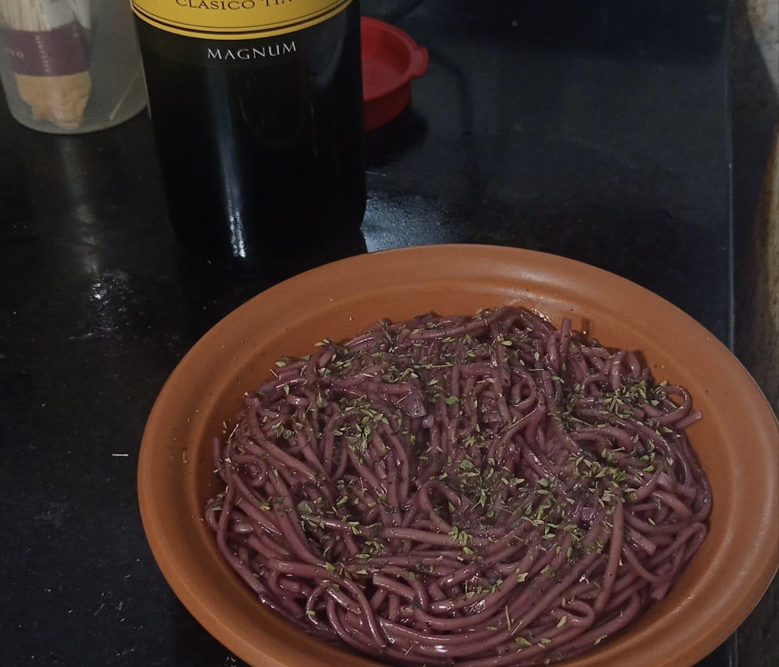 A terracotta bowl filled with spaghetti coated in a purple sauce, topped with herbs. A bottle of red wine is visible in the background on a dark countertop.