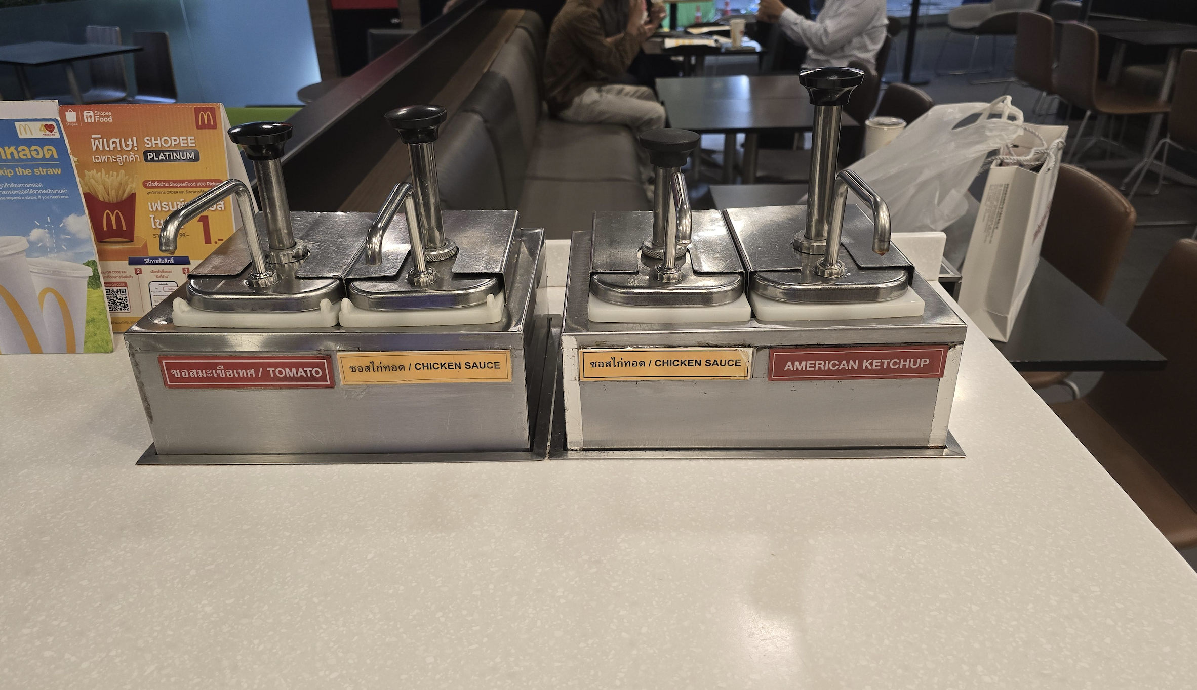 Two condiment dispensers on a counter, labeled for tomato sauce, chicken sauce, and American ketchup, in a fast food restaurant. Tables and chairs are visible in the background.
