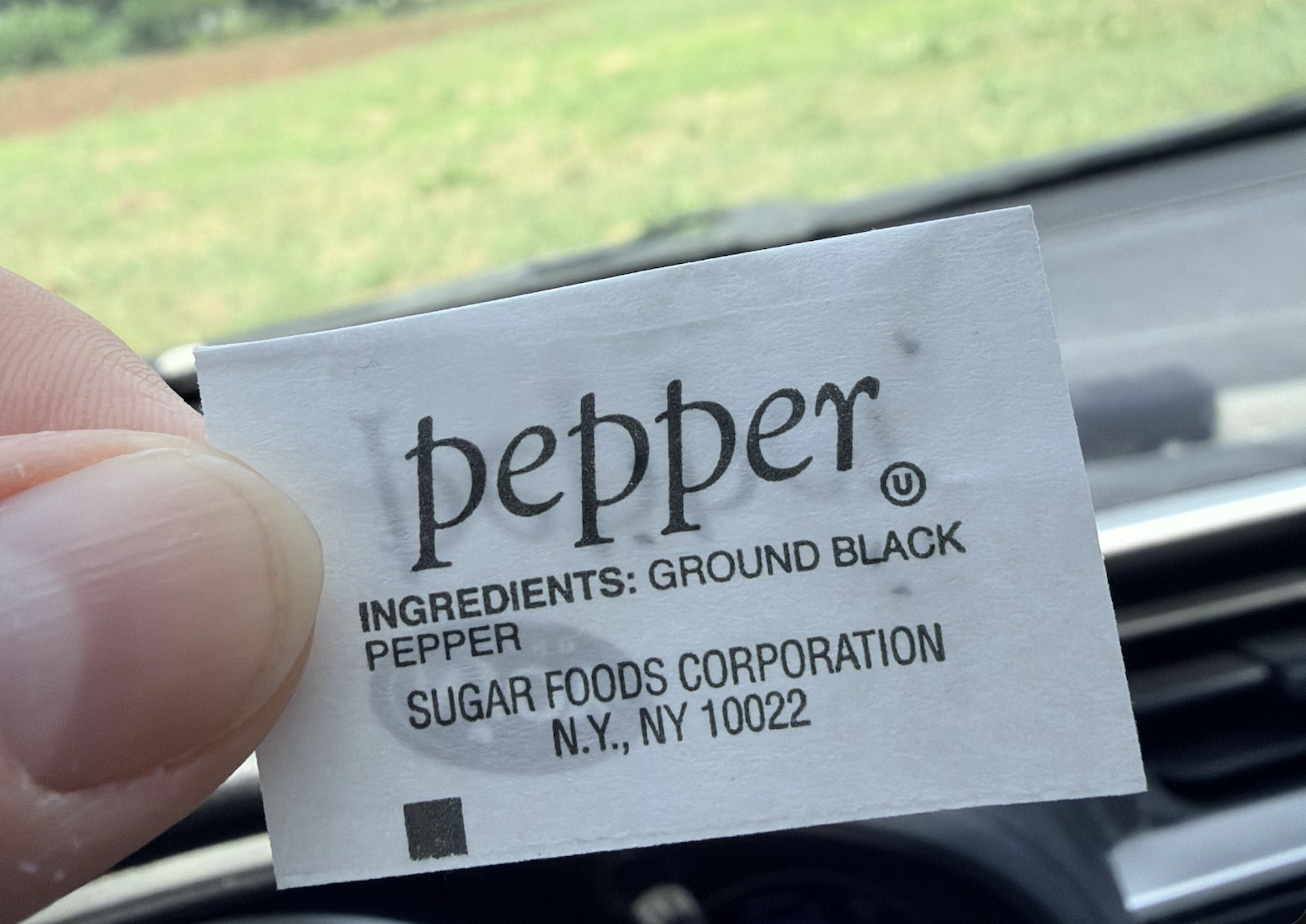 A hand holds a small white packet labeled “pepper,” listing ground black pepper as the ingredient, with the manufacturer Sugar Foods Corporation and a New York address below. A blurred car dashboard and grassy field are in the background.