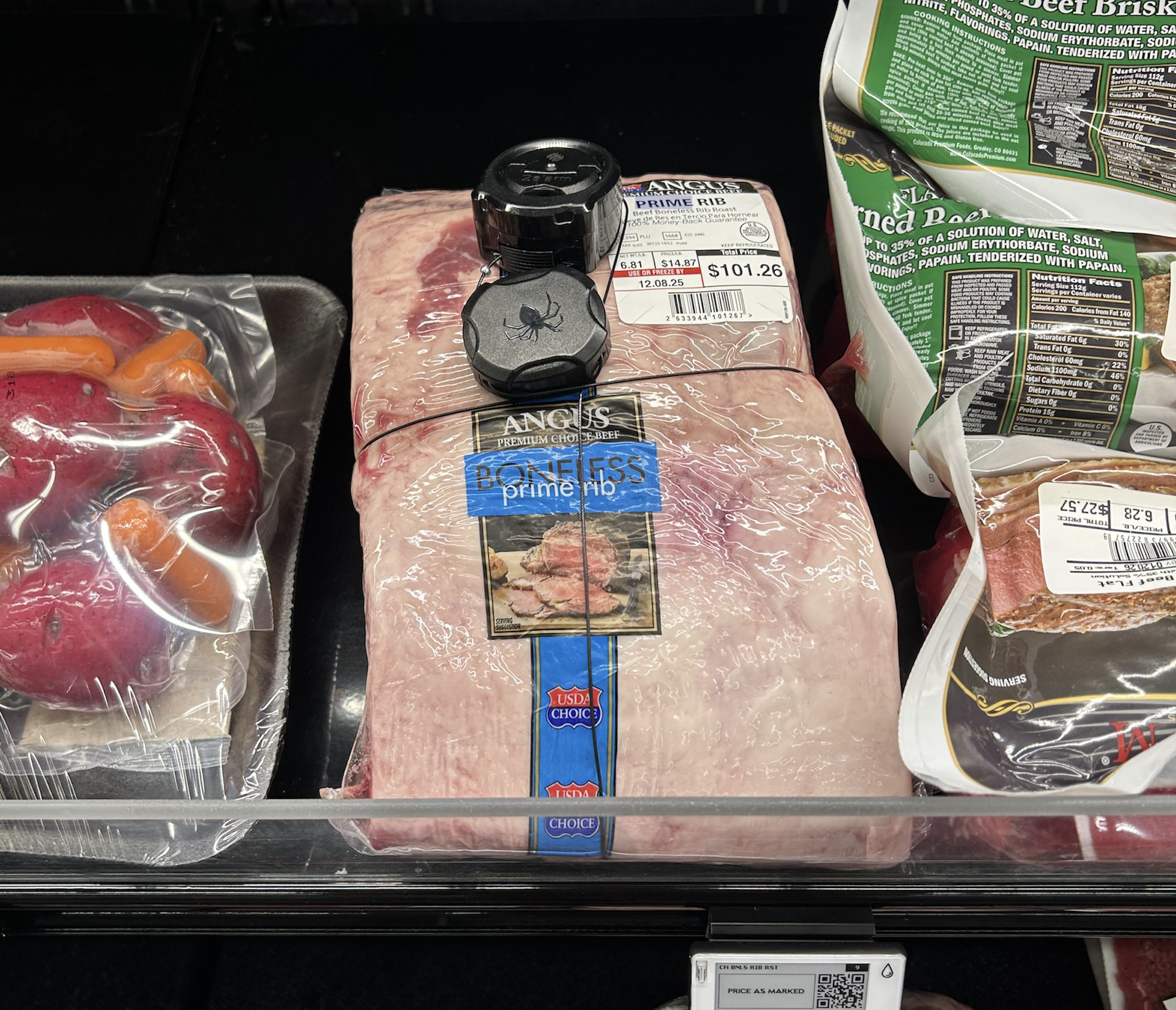 A boneless Angus prime rib roast in plastic wrap with a security tag and price label, displayed in a grocery meat case between packaged beef and other meat products.