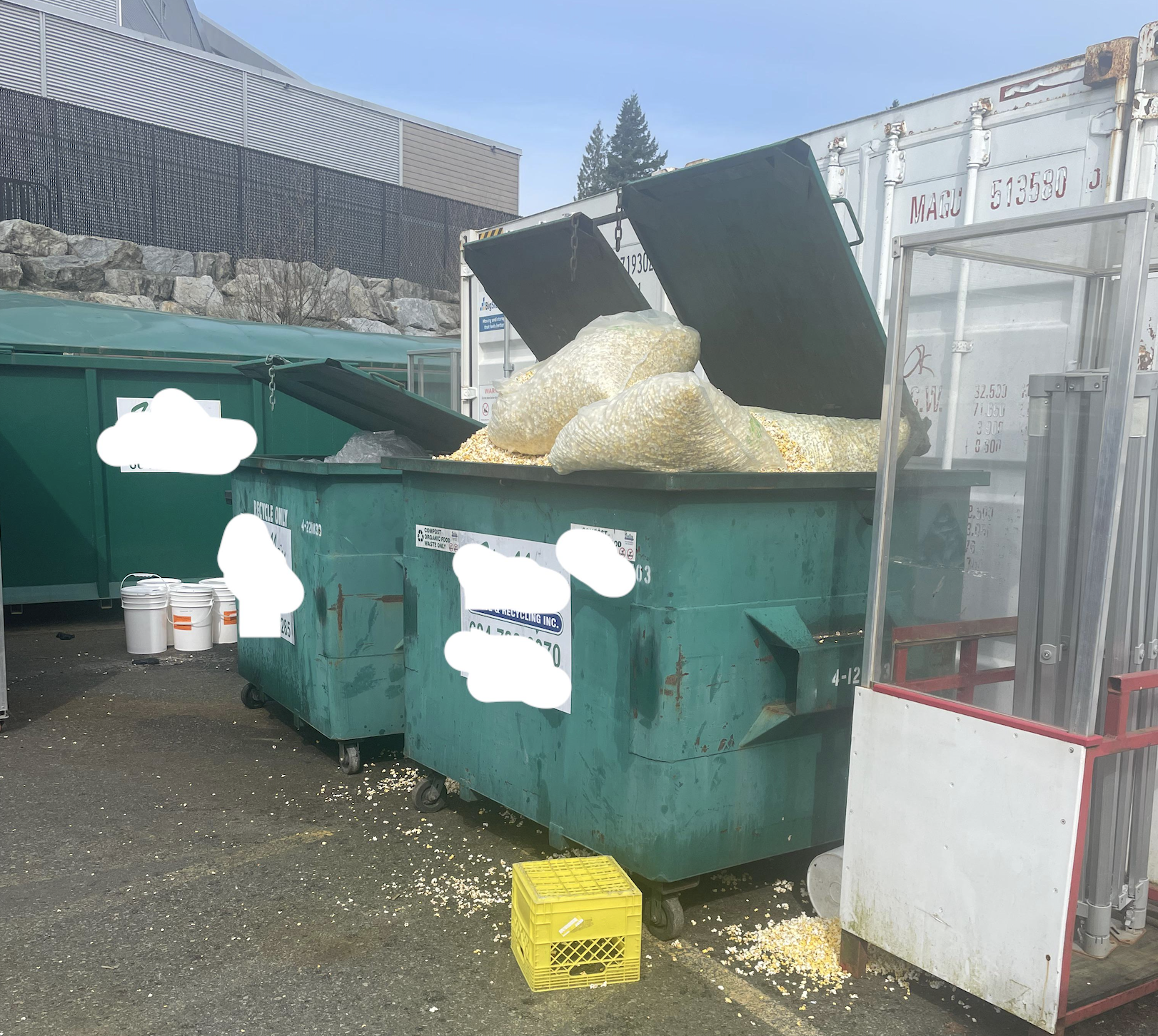 Two large green dumpsters overflowing with bags of popcorn, with popcorn scattered on the ground nearby. White buckets and a yellow crate are next to the dumpsters. Industrial containers and buildings are in the background.