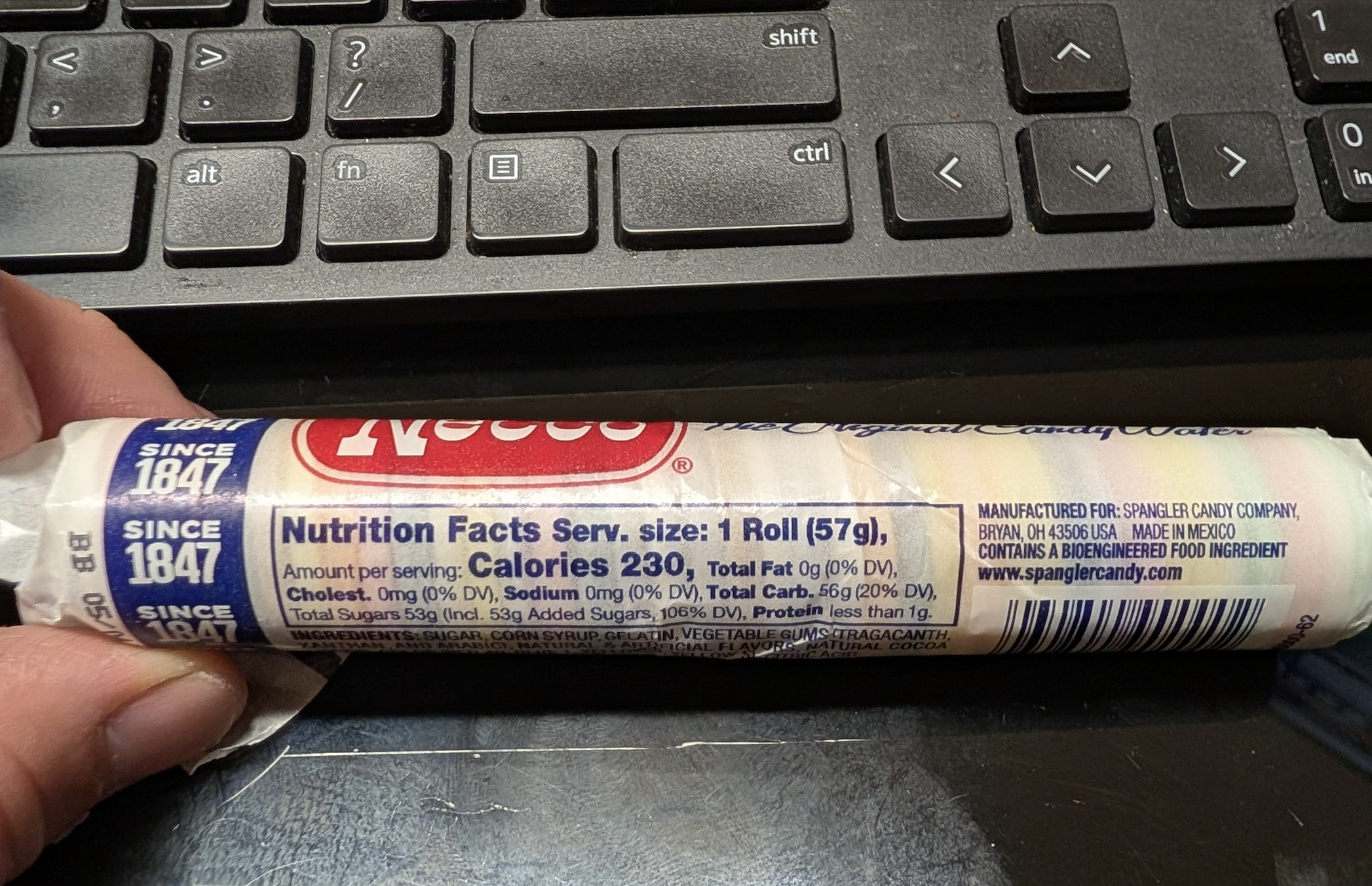 A hand holds a roll of Necco wafers showing nutrition facts and ingredients, next to a black computer keyboard.