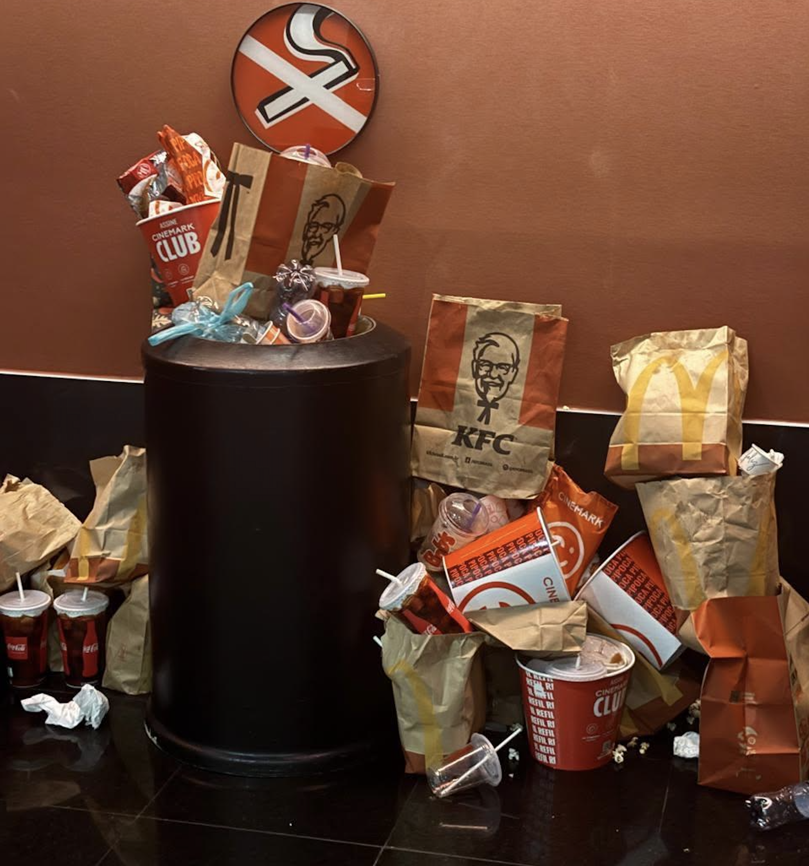 An overflowing trash can surrounded by fast food bags, cups, and containers from KFC and McDonald’s under a “No Smoking” sign.