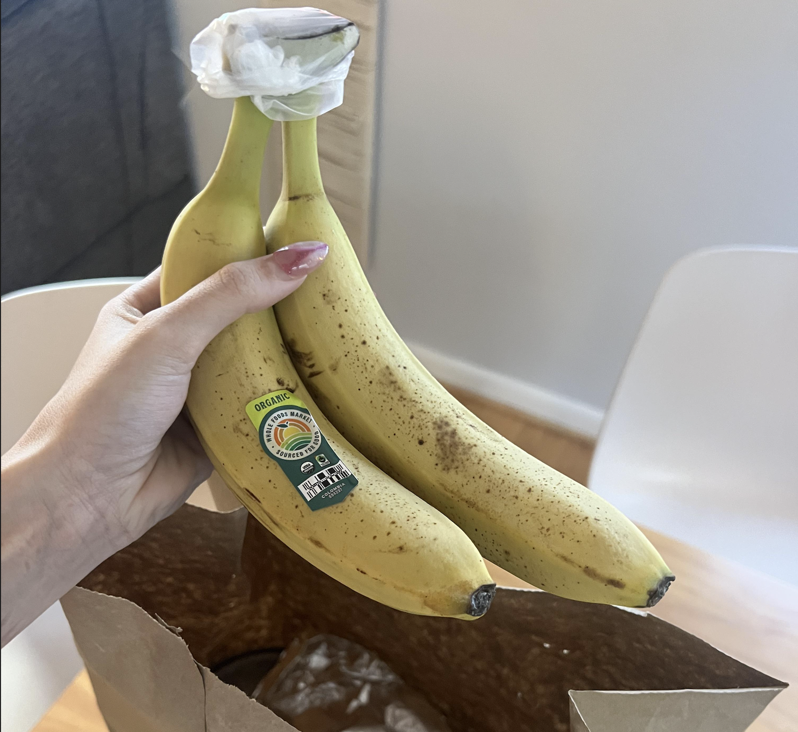 A hand holding two ripe bananas with stickers, above a brown paper bag. The background includes a white chair and a wooden floor.