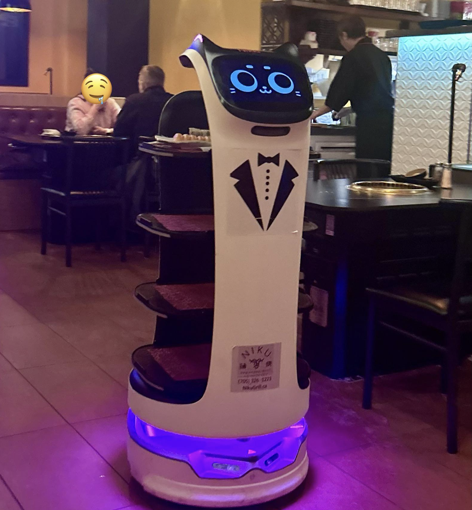 A cat-themed robot waiter with shelves and a tuxedo design serves at a restaurant, while diners and a staff member are in the background.