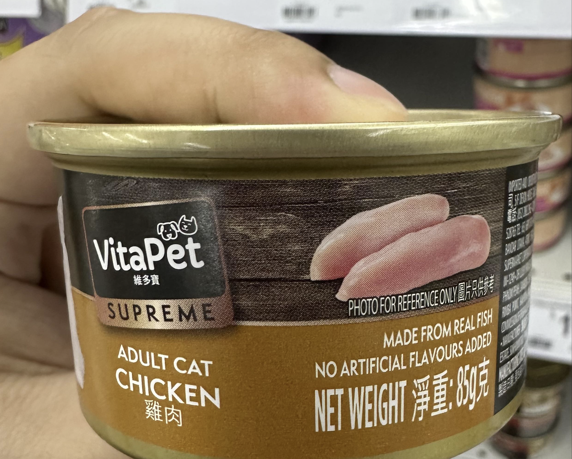 A hand holds a can of VitaPet Supreme adult cat food, chicken flavor. The label shows slices of chicken, highlights “made from real fish,” and has a net weight of 85g.
