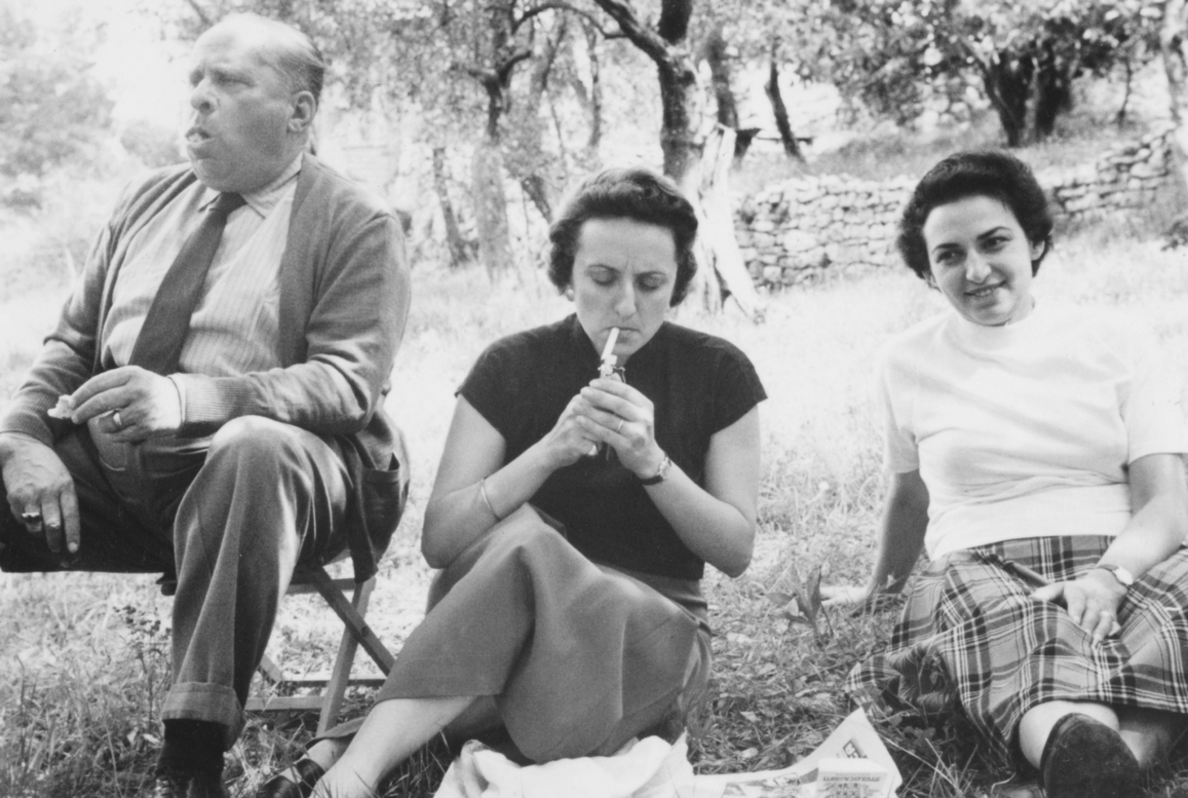 Three adults sit on grass outdoors. The man on the left wears a suit, the woman in the middle drinks from a bottle, and the woman on the right smiles. There are trees and stone walls in the background.