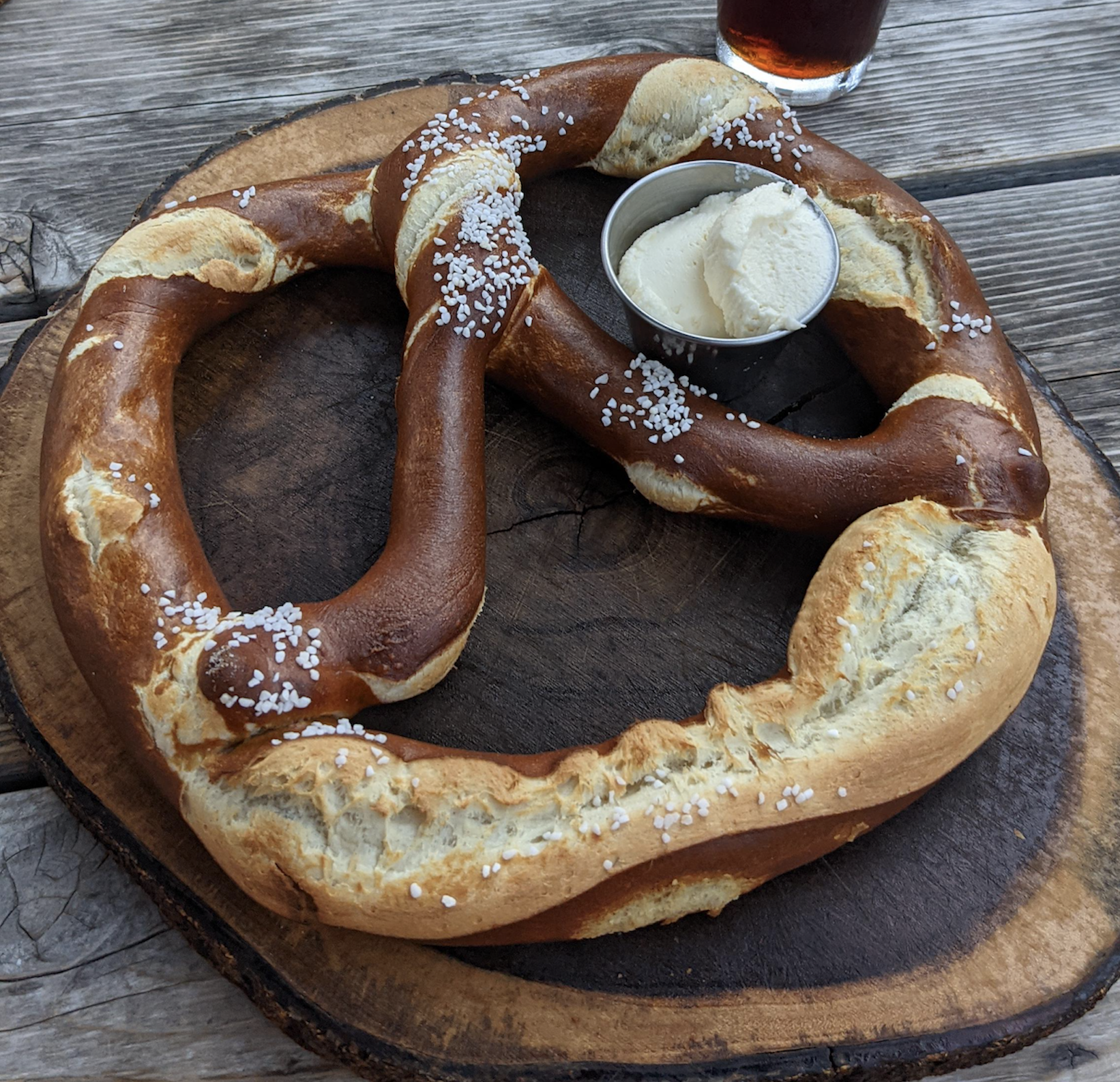 A large soft pretzel topped with coarse salt sits on a wooden board, served with a small metal cup of creamy white dipping sauce. A glass of dark beverage is partly visible in the background.