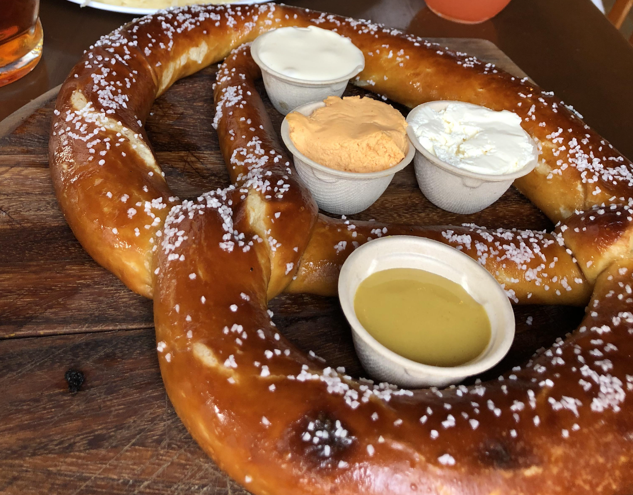 A large, soft pretzel topped with coarse salt is served on a wooden board with four dipping sauces: white, orange, and yellow spreads in small cups.
