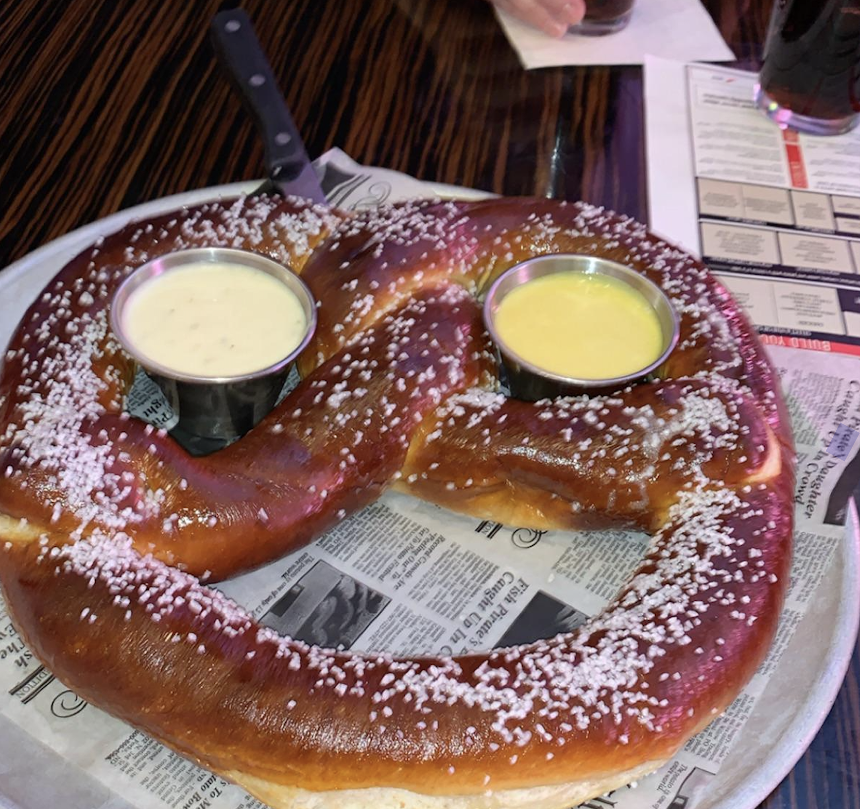 A large salted pretzel on a tray, served with two dipping sauces in cups placed on the pretzel, one white and one yellow, with a knife resting on top. The tray is lined with newspaper-style paper.