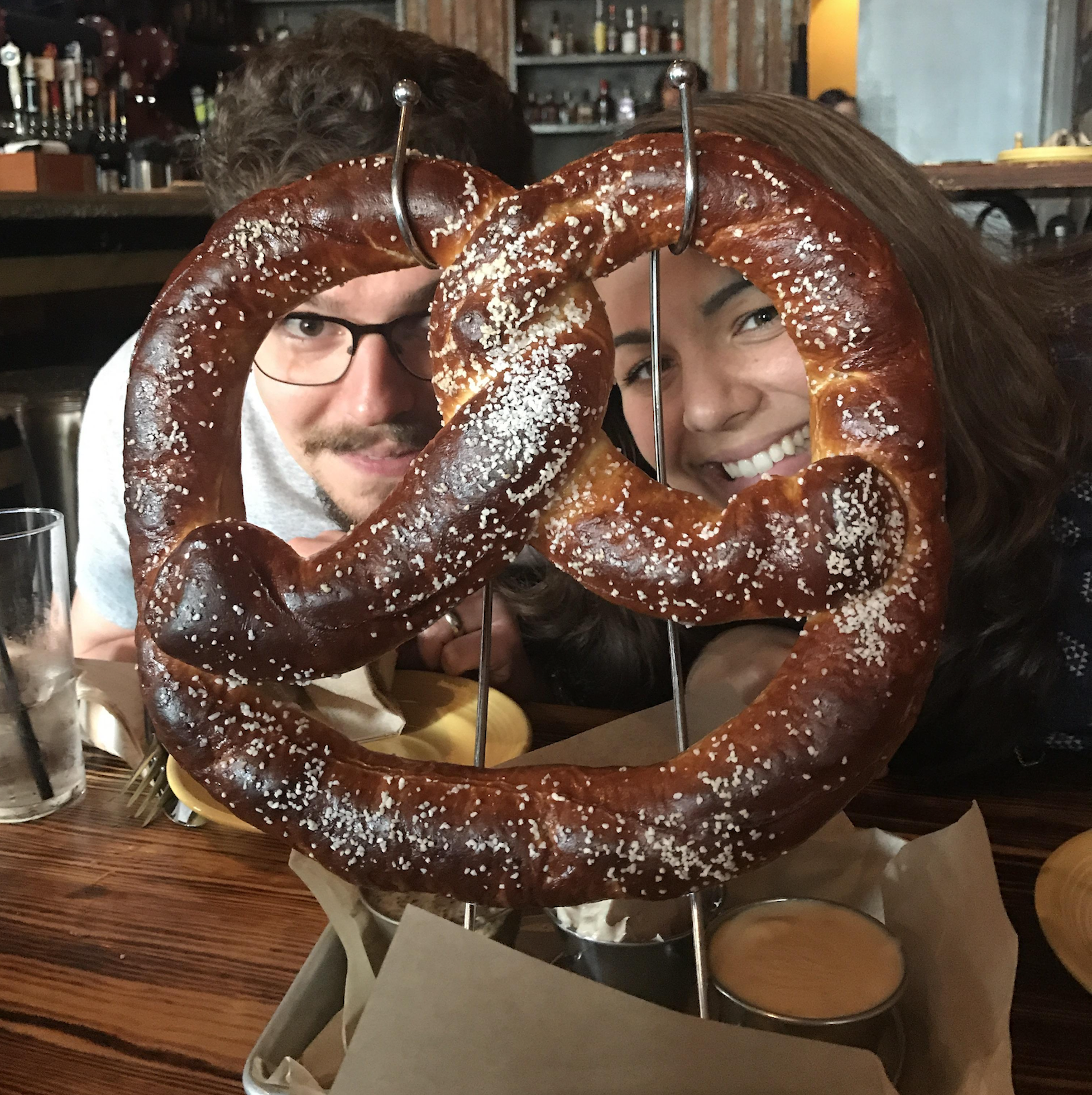 Two people smile behind a large, salted pretzel on a stand at a restaurant table, with dipping sauces and drinks nearby.