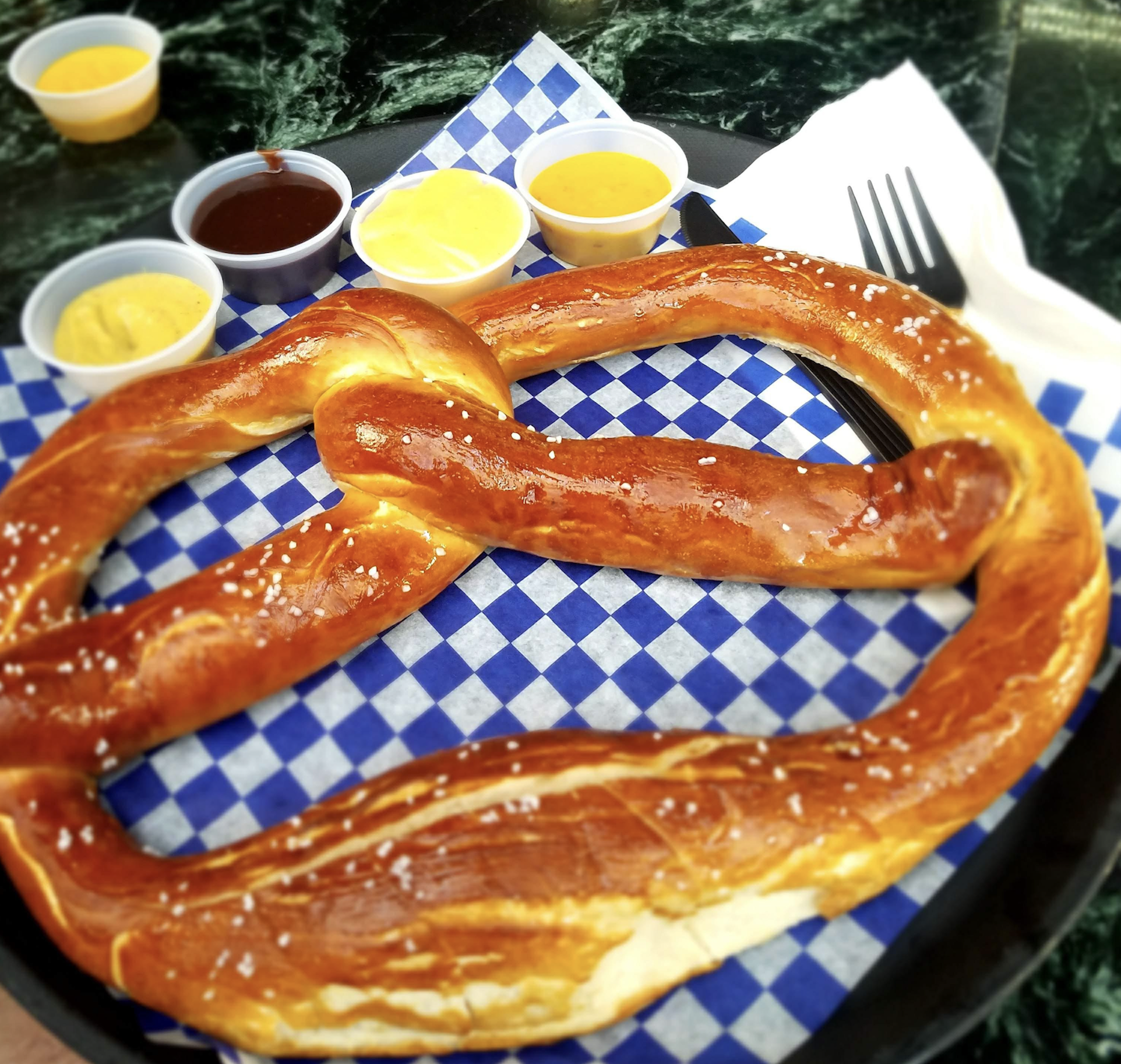 A large soft pretzel topped with salt sits on blue-and-white checkered paper, served with four dipping sauces, a fork, and napkins on a dark green marble table.