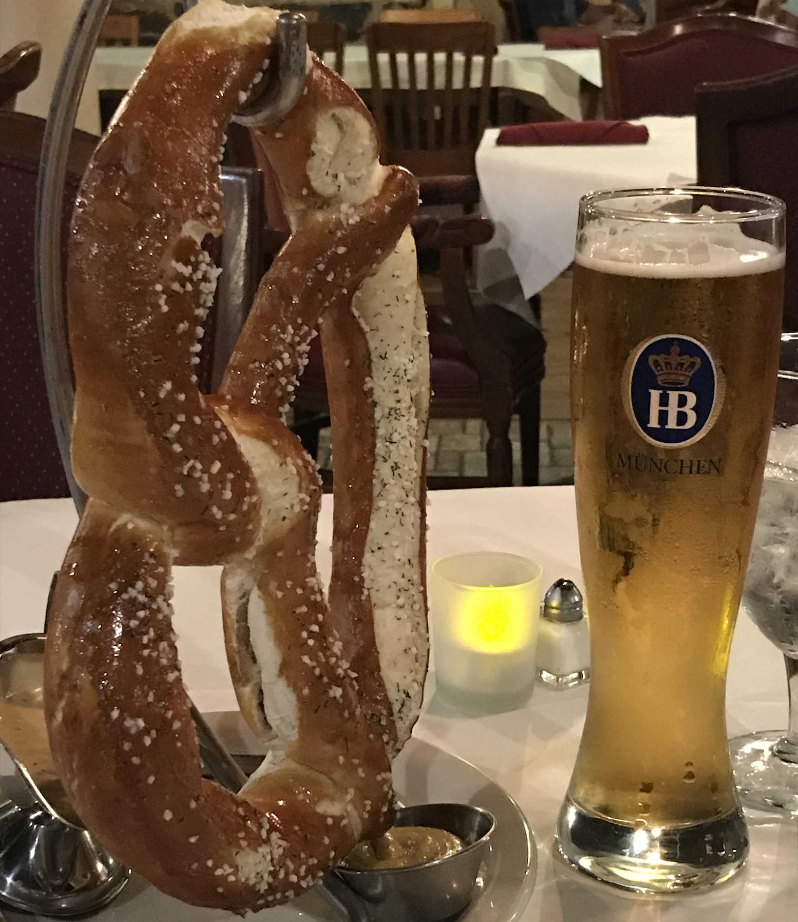 A large soft pretzel on a metal stand with dipping sauces next to a tall glass of beer labeled “HB München” on a restaurant table.