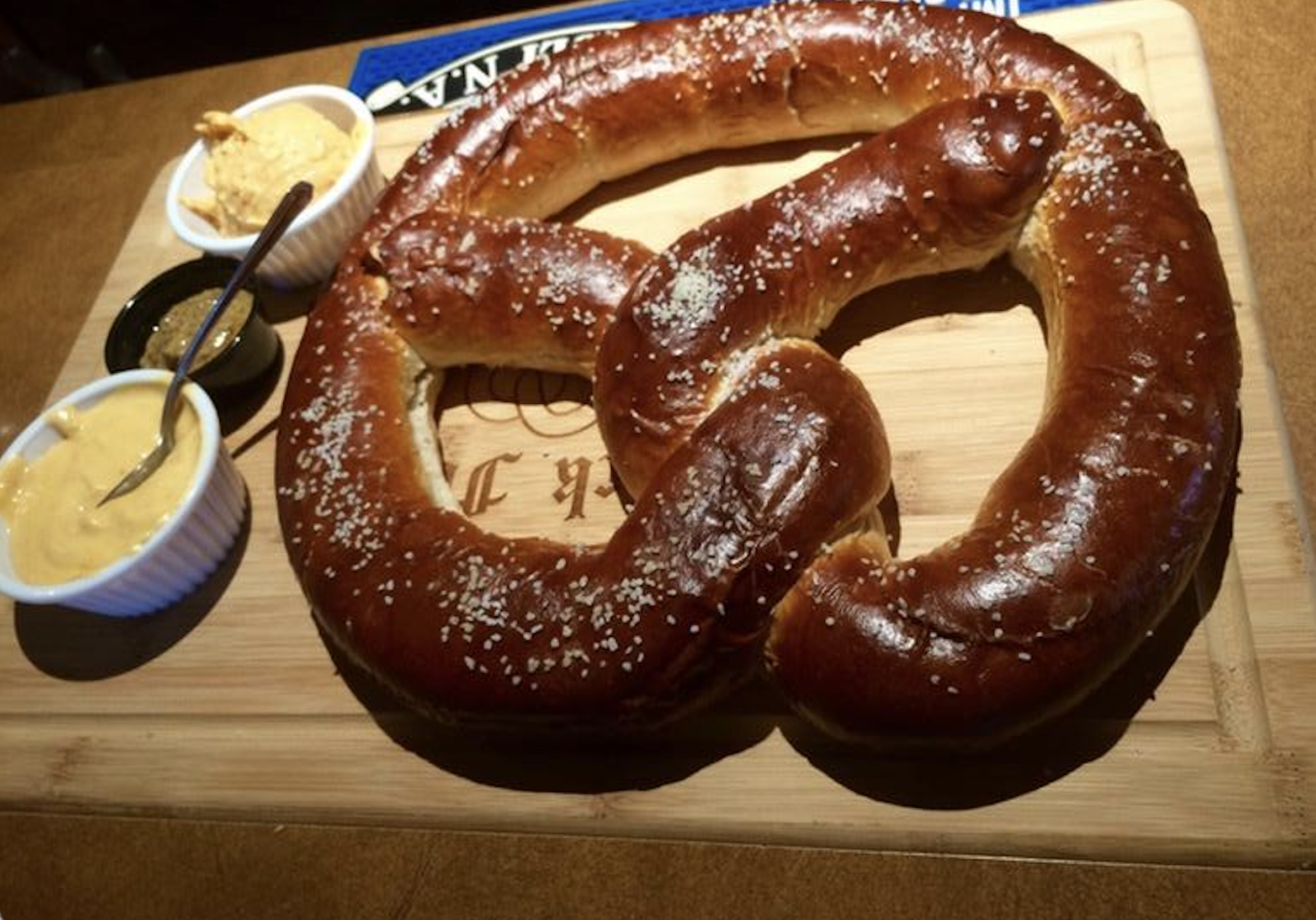A large, salted soft pretzel on a wooden board, served with three small bowls containing different dipping sauces.