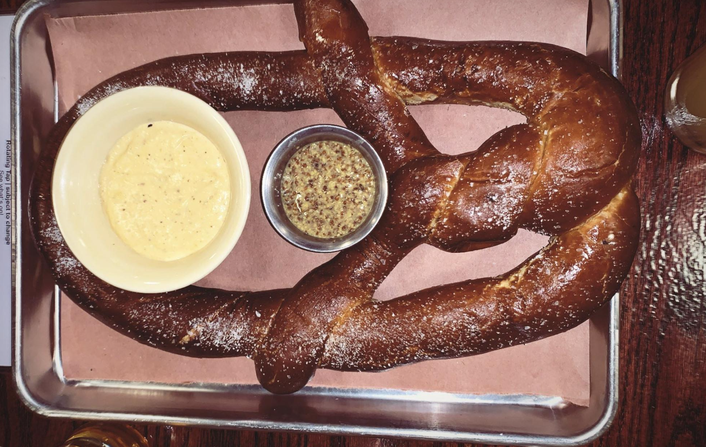 A large salted pretzel on a metal tray with a side of cheese dip in a bowl and whole grain mustard in a small metal cup.