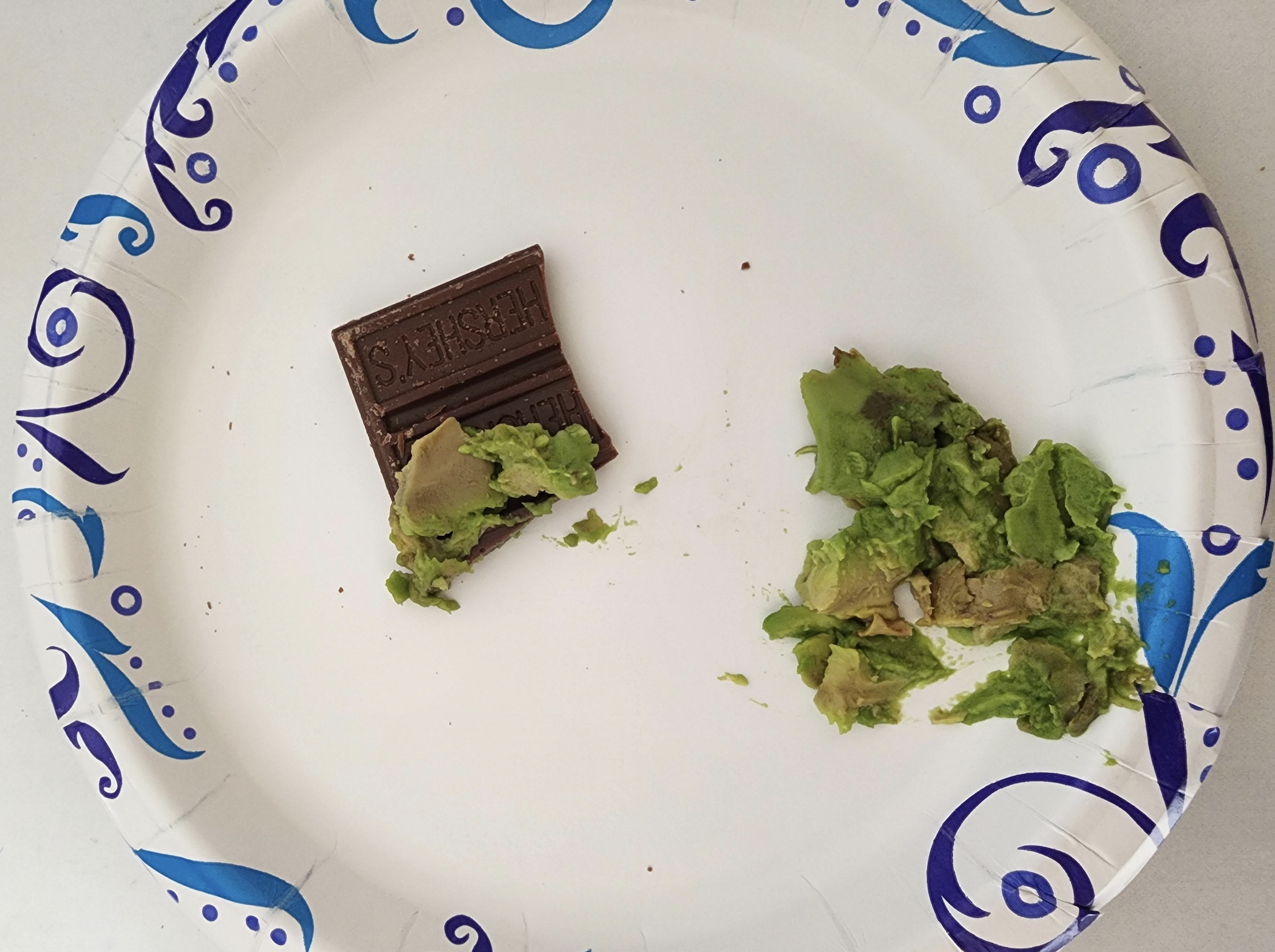 A paper plate with one piece of chocolate topped with mashed avocado on the left and a small pile of mashed avocado on the right.