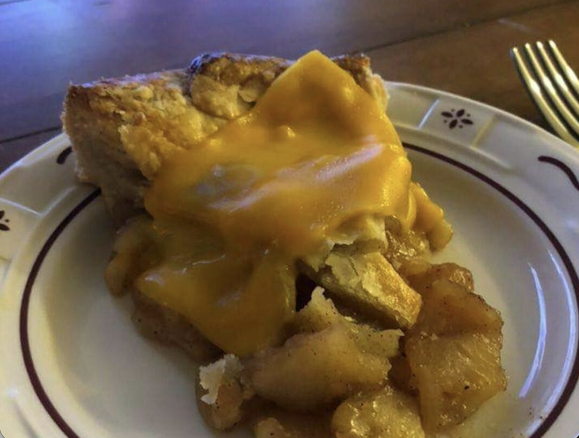 A slice of apple pie topped with a melted slice of cheddar cheese sits on a decorative plate next to a fork on a wooden table.