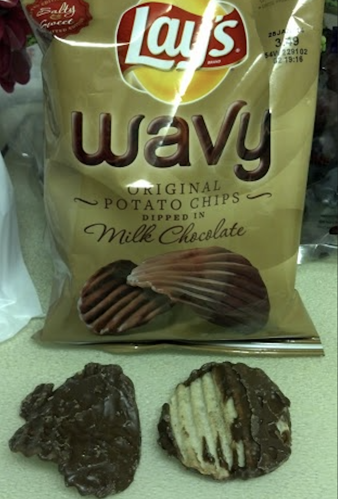 A bag of Lay’s Wavy Original Potato Chips dipped in milk chocolate is shown with two chocolate-covered wavy chips in front of the bag on a light surface.