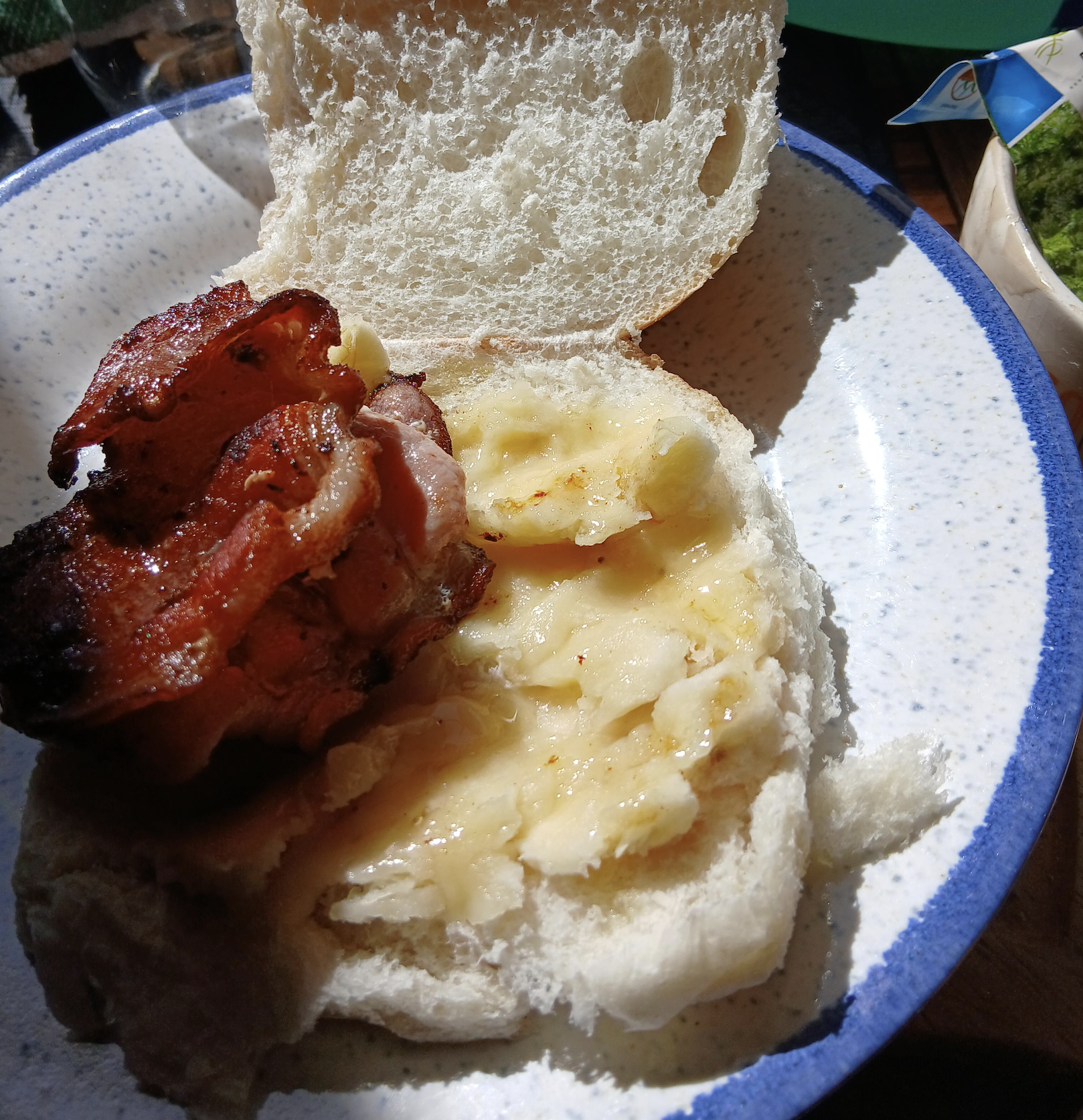 A sandwich bun with mashed banana and crispy bacon inside, shown on a white speckled plate in bright sunlight.