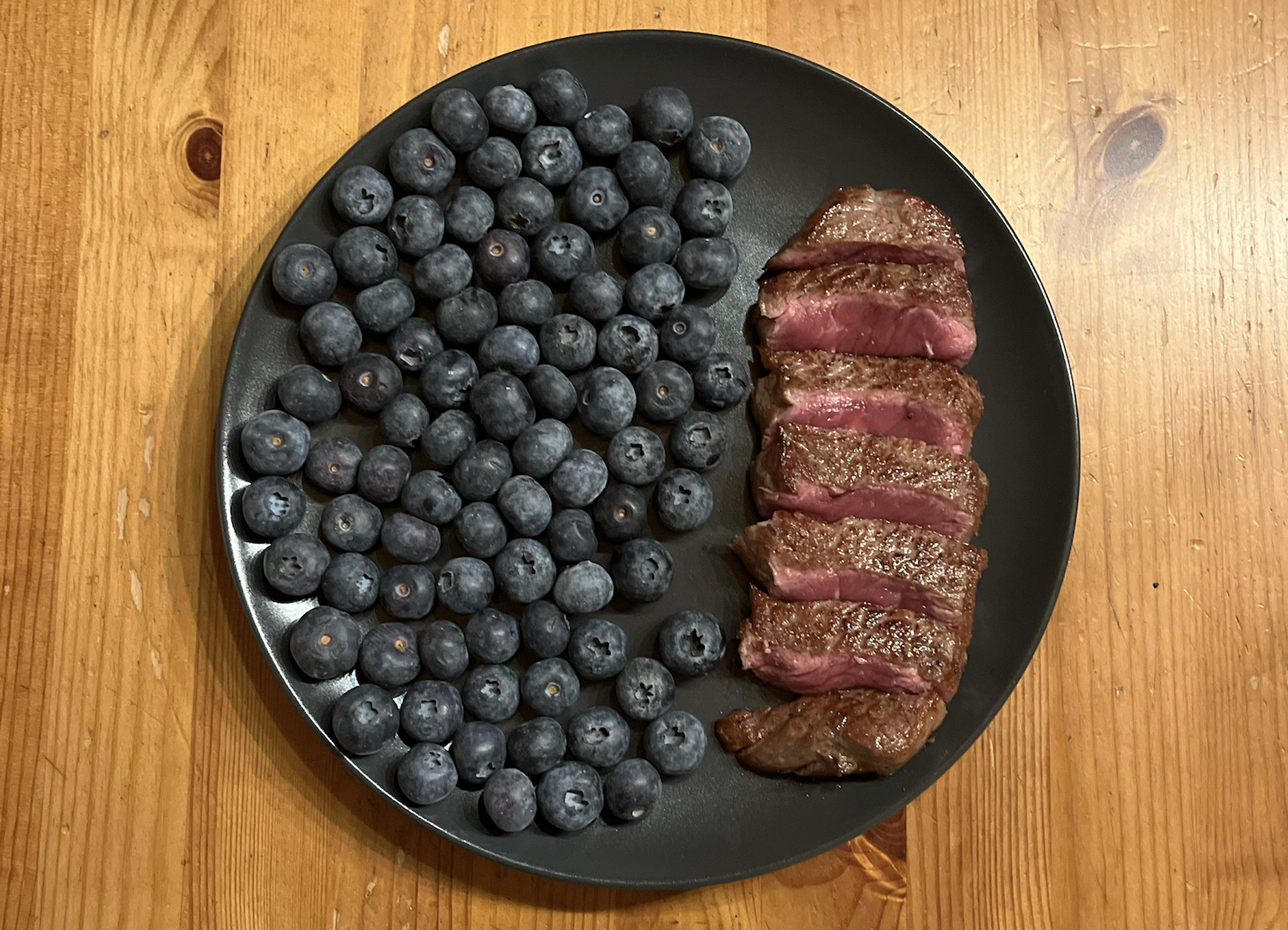 A black plate on a wooden table holds a pile of fresh blueberries on the left and several slices of medium-rare steak on the right.