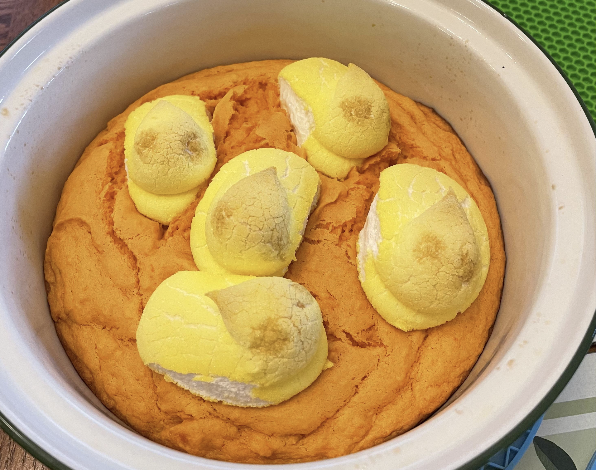 A round orange cake in a white dish, topped with five semi-melted yellow marshmallow chicks.
