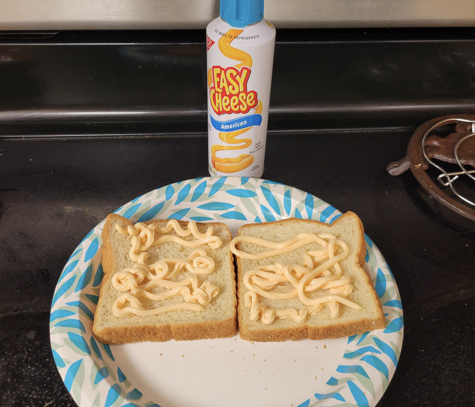 Two slices of bread on a paper plate, each topped with squiggles of Easy Cheese. The Easy Cheese can is behind the plate on a kitchen stove.