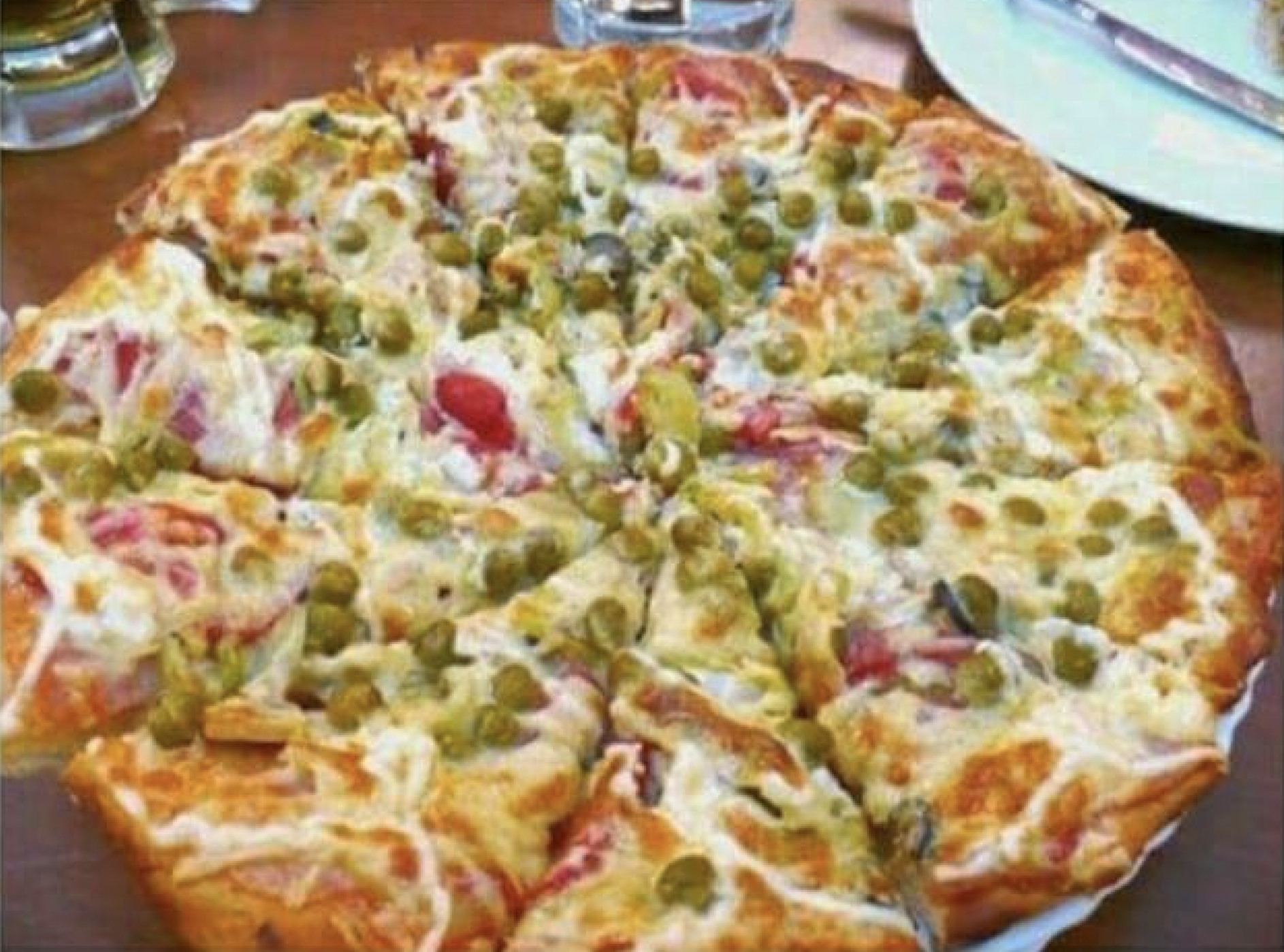 A baked pizza topped with green peas, melted cheese, and pieces of tomato, sliced into eight pieces, placed on a wooden table near a plate and utensils.